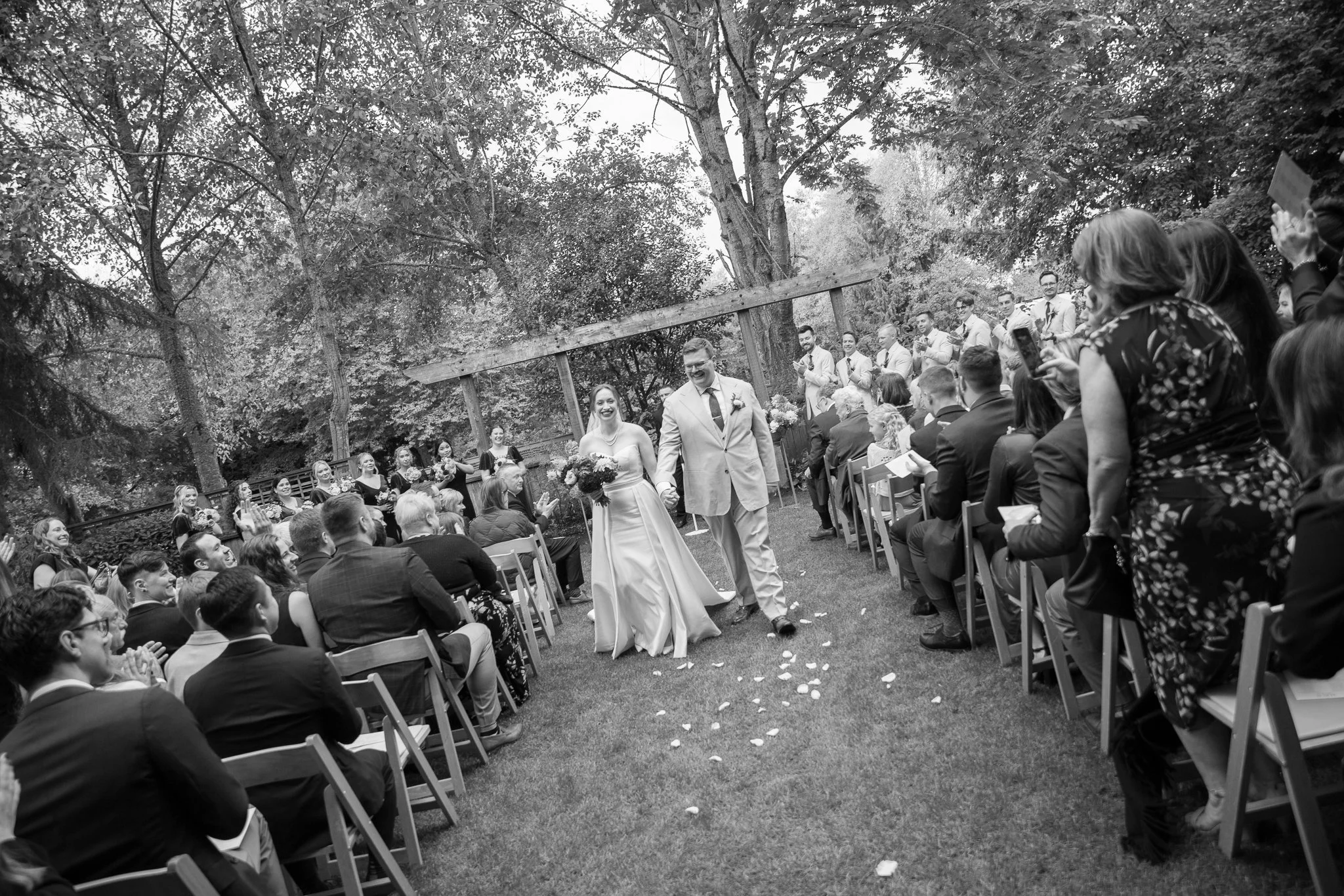 A black and white photo of a wedding ceremony outdoors, with a bride and groom walking hand in hand down the aisle. The bride is wearing a wedding dress and holding a bouquet, and the groom is in a light-colored suit. Guests are seated on either side