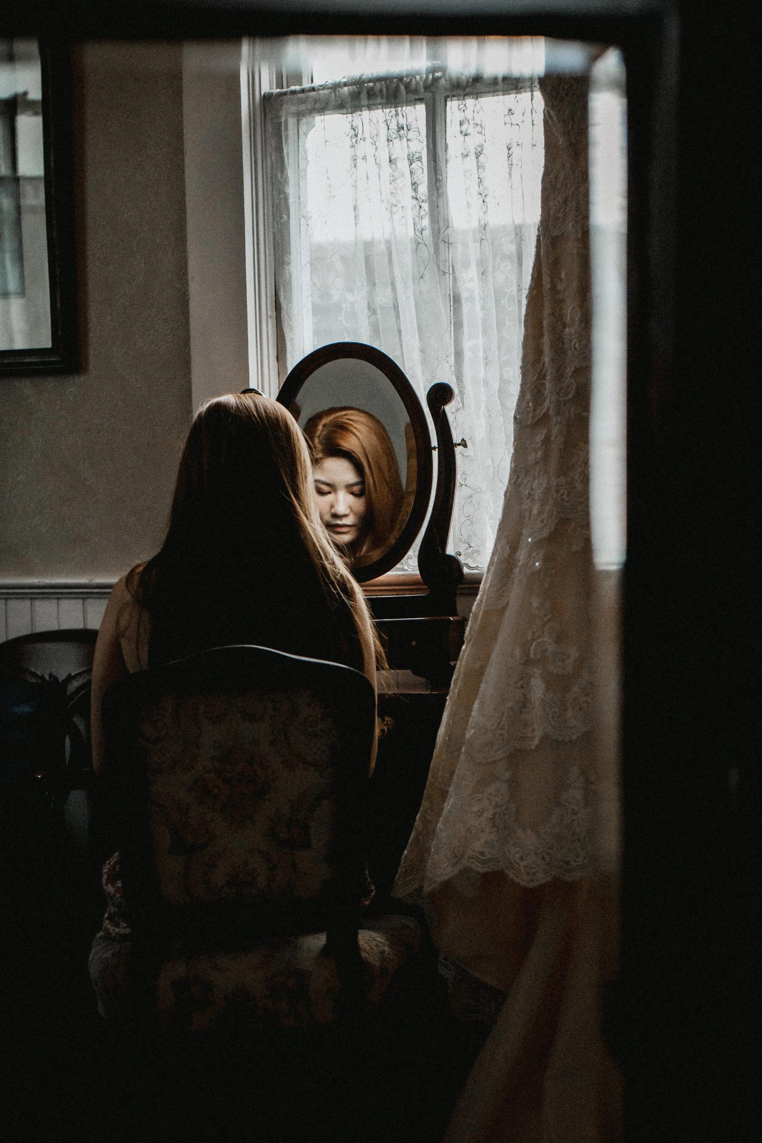 Woman sitting on the floor in front of a mirror in a dimly lit room, with a lace curtain partially covering the window.