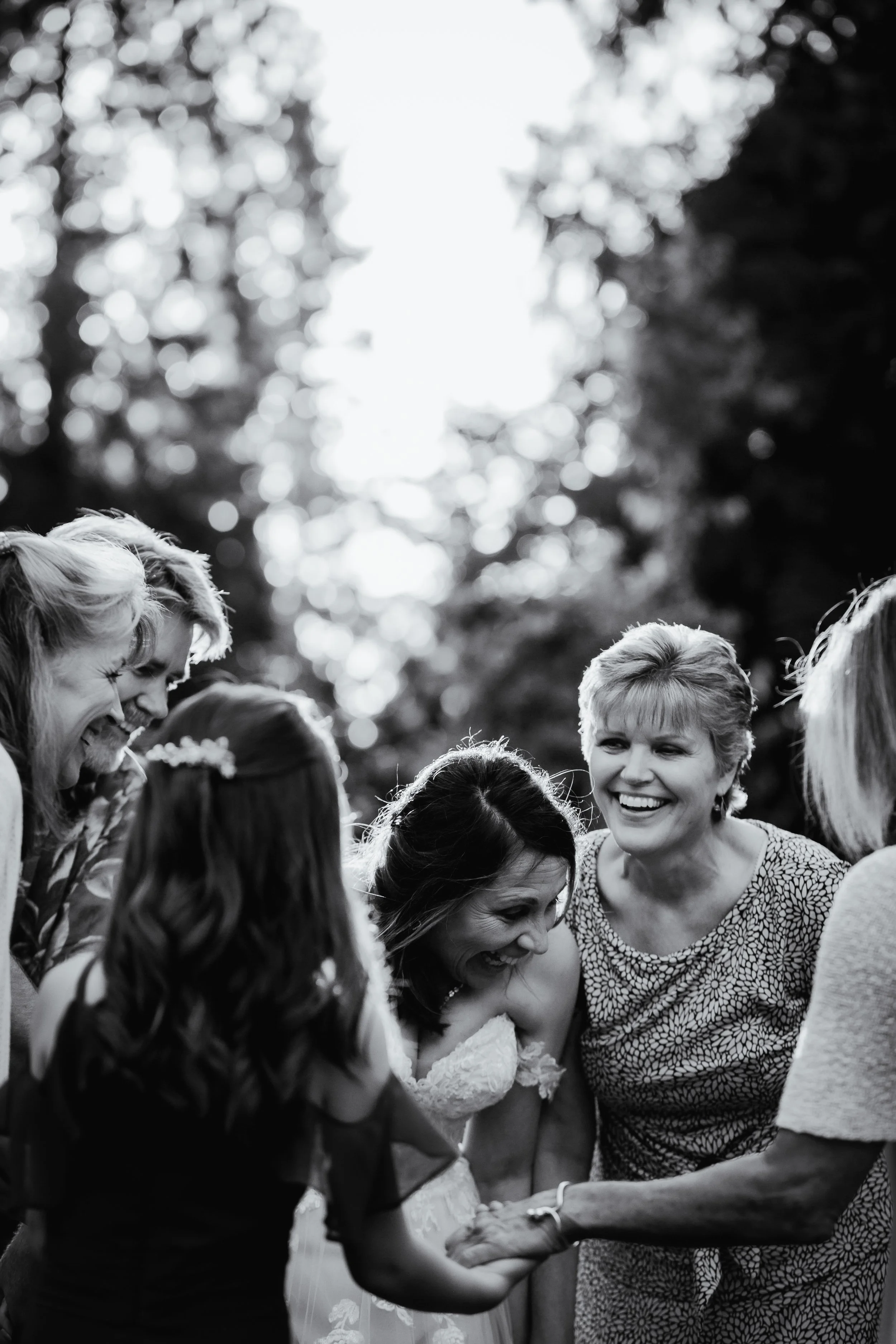 A group of women and a girl are smiling and gathered in a circle outdoors, holding hands, during a joyful moment at a wedding.