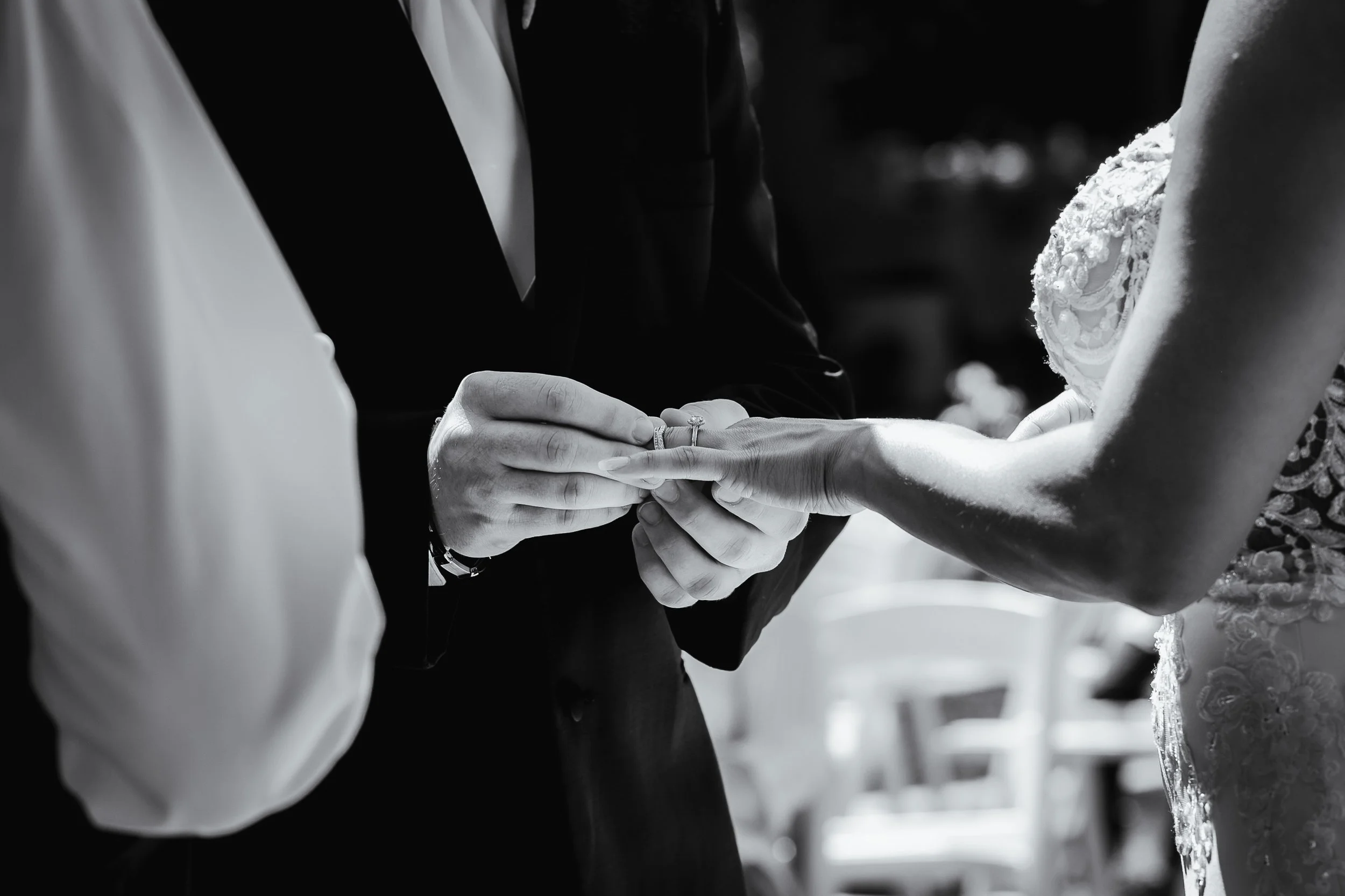 A close-up black-and-white photo of a groom placing a wedding ring on the bride's finger, with the bride wearing a lace wedding dress.