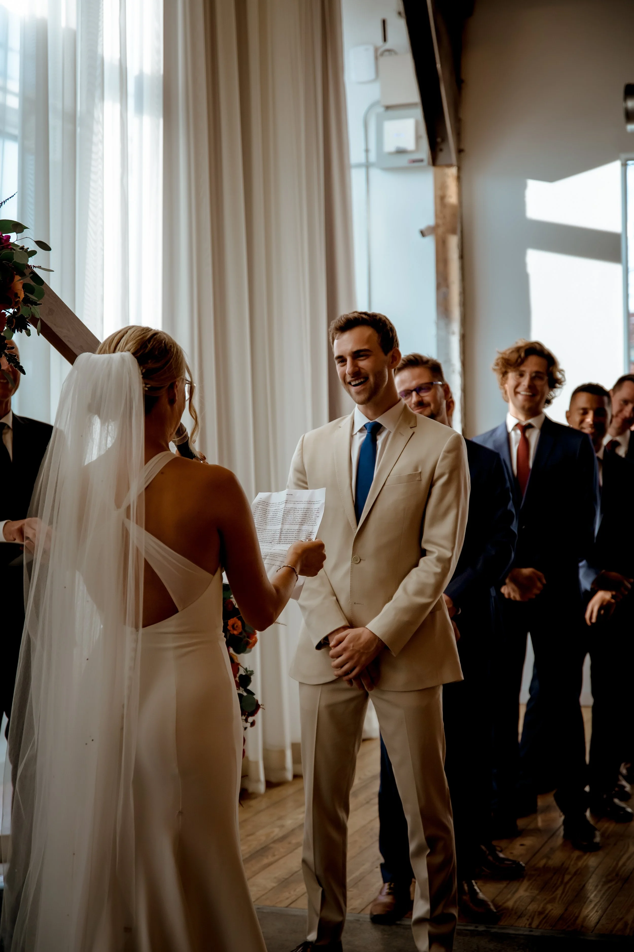 A wedding ceremony with a woman in a white dress and veil, reading vows to a man in a beige suit, with guests in formal attire smiling in the background.