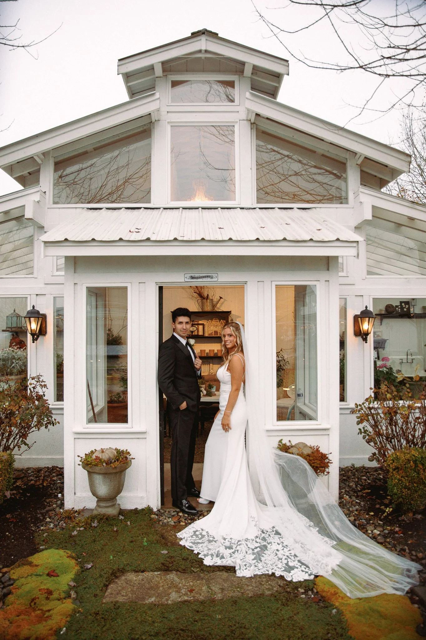 A bride and groom standing at the entrance of a white greenhouse-style house, with the bride in a white wedding dress and veil, and the groom in a black tuxedo.