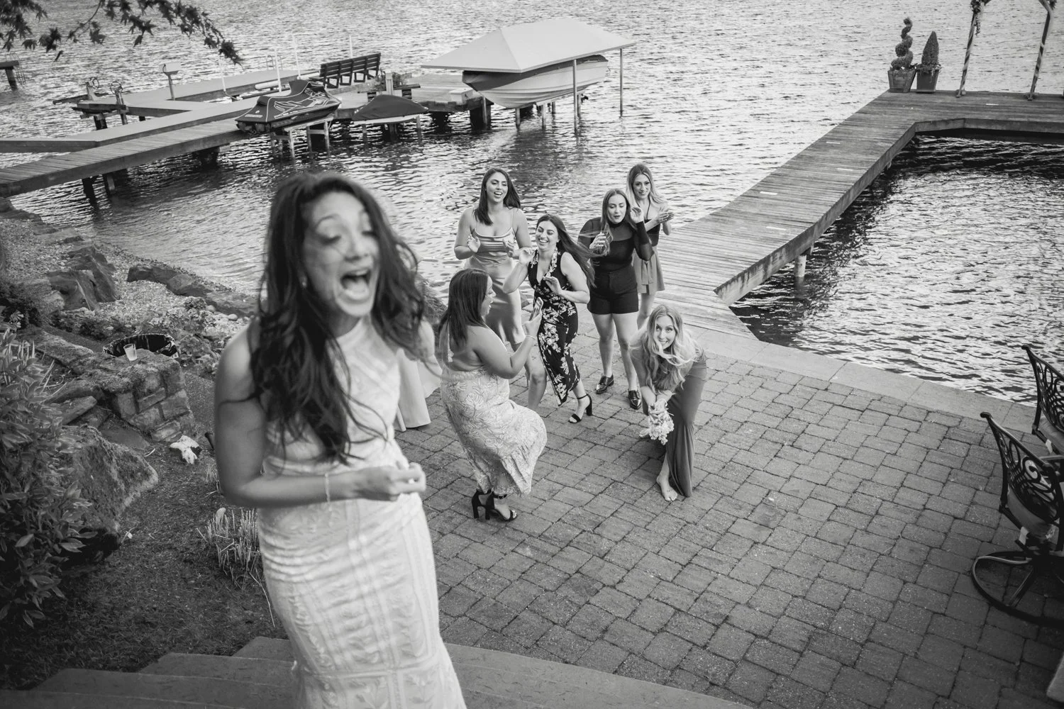 A group of women at a waterfront venue, with one woman in the foreground laughing and the others in the background celebrating near a dock with boats, holding drinks, and a woman with a bouquet.