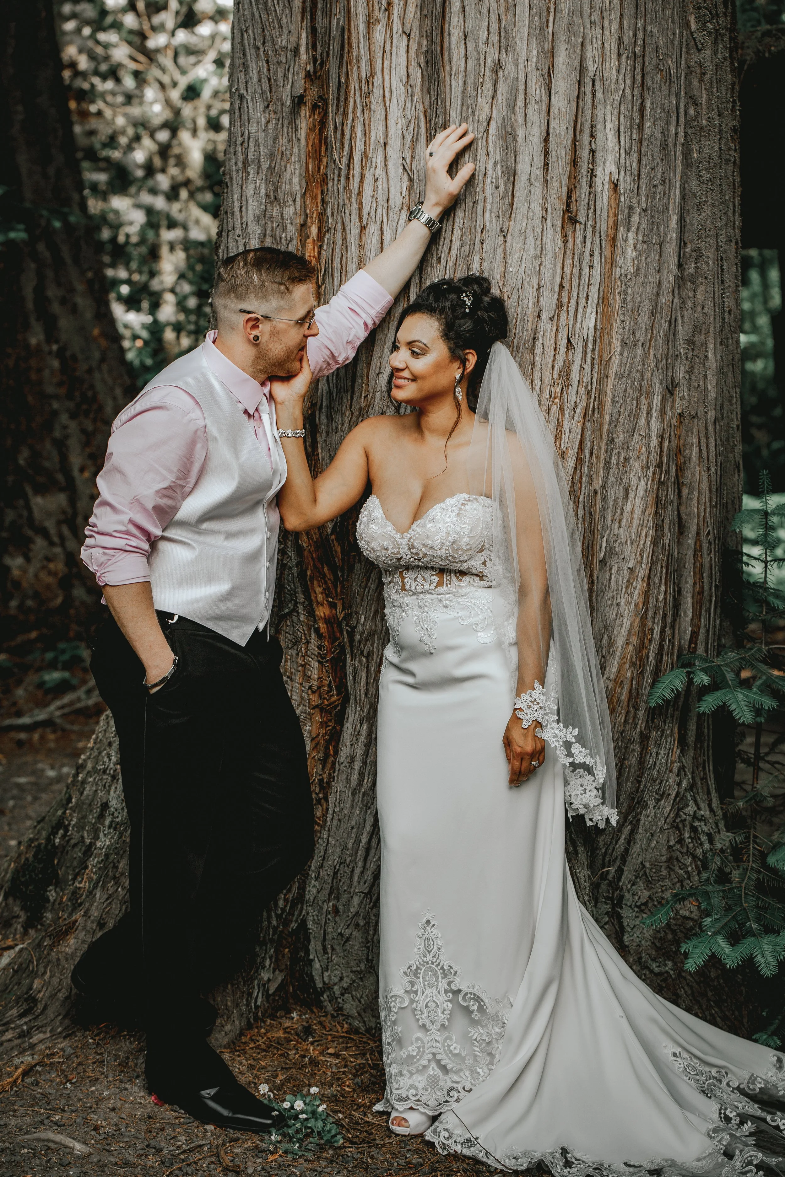A newlywed couple poses outdoors in front of a large tree, with the bride wearing a white lace wedding gown and veil, and the groom dressed in a light pink shirt and vest, as they look at each other romantically.