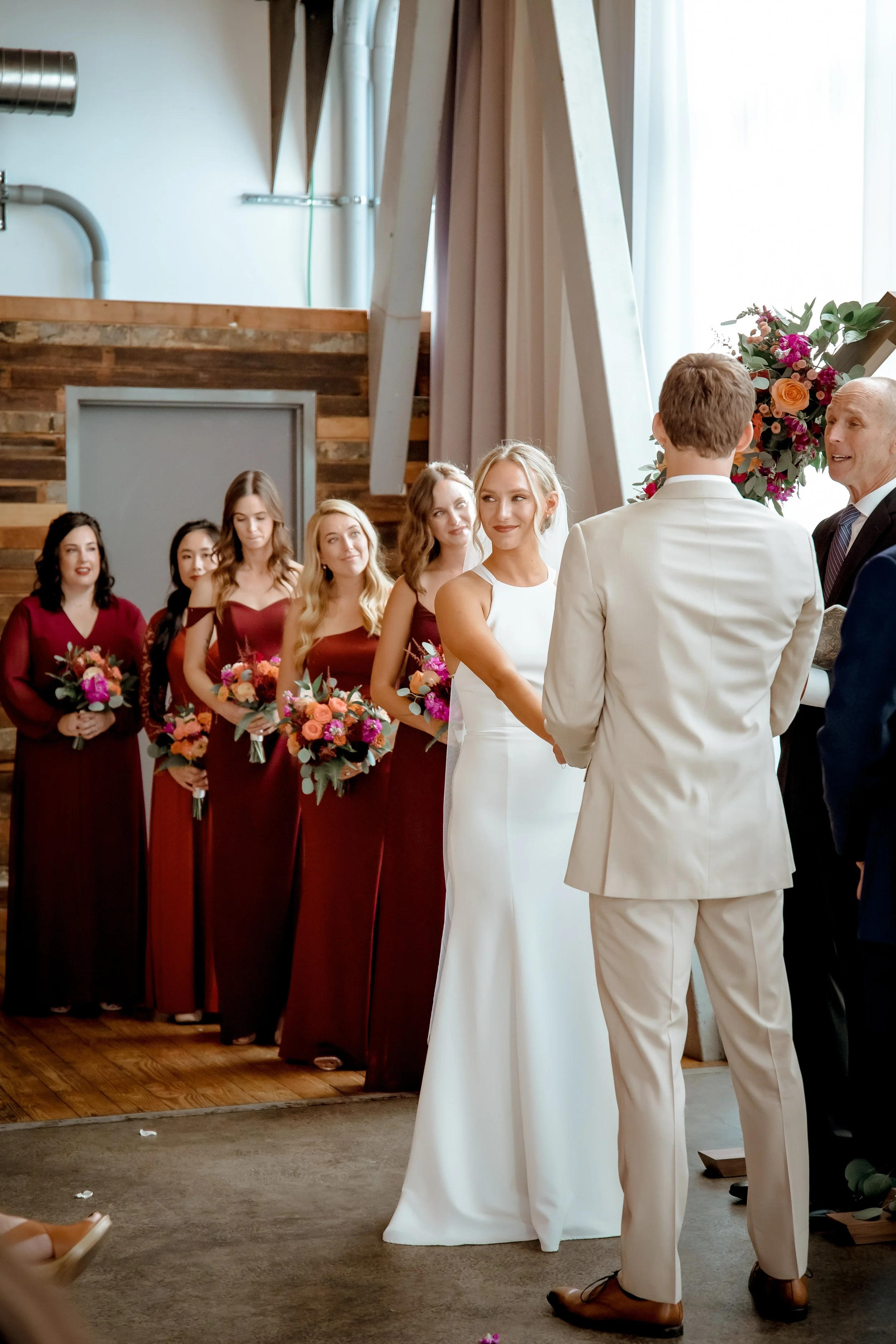 A wedding ceremony with a bride and groom standing in front of a wedding party, which includes bridesmaids in burgundy dresses holding bouquets, and an officiant speaking to the couple, with a floral arrangement nearby.