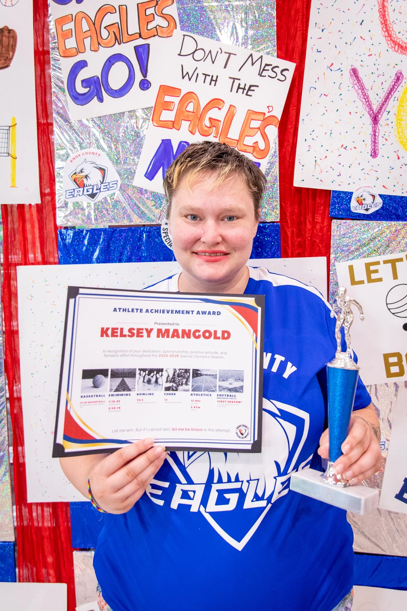 portrait of a person standing in front of a special olympics back drop, holding their trophy and certificate