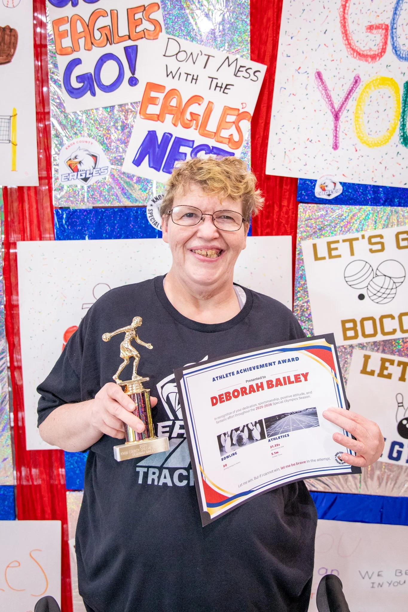 portrait of a person standing in front of a special olympics back drop, holding their trophy and certificate
