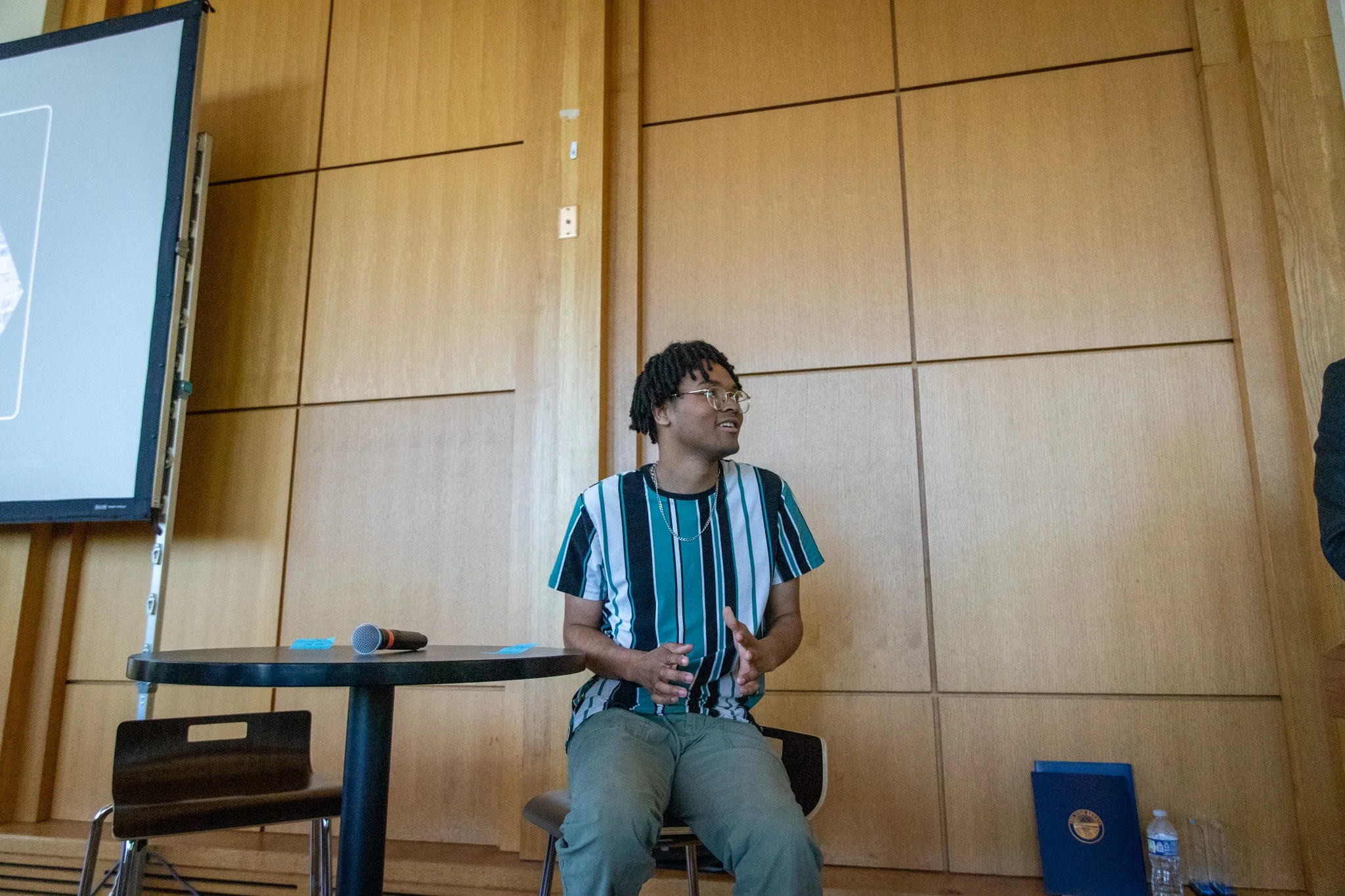 Person sits beside a small table, gesturing while speaking in a wood-paneled event space with a microphone resting nearby.