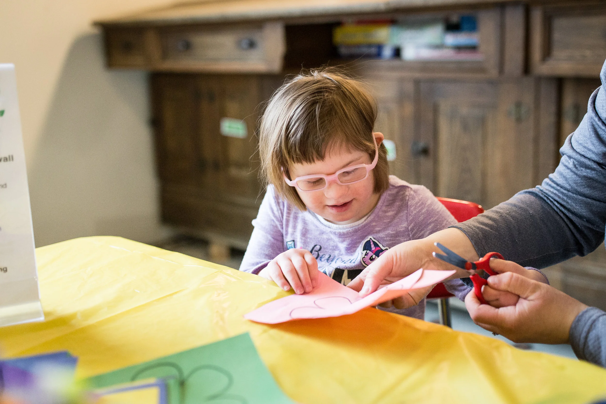 little girl with Down Syndrome cutting a piece of paper with help from her mom