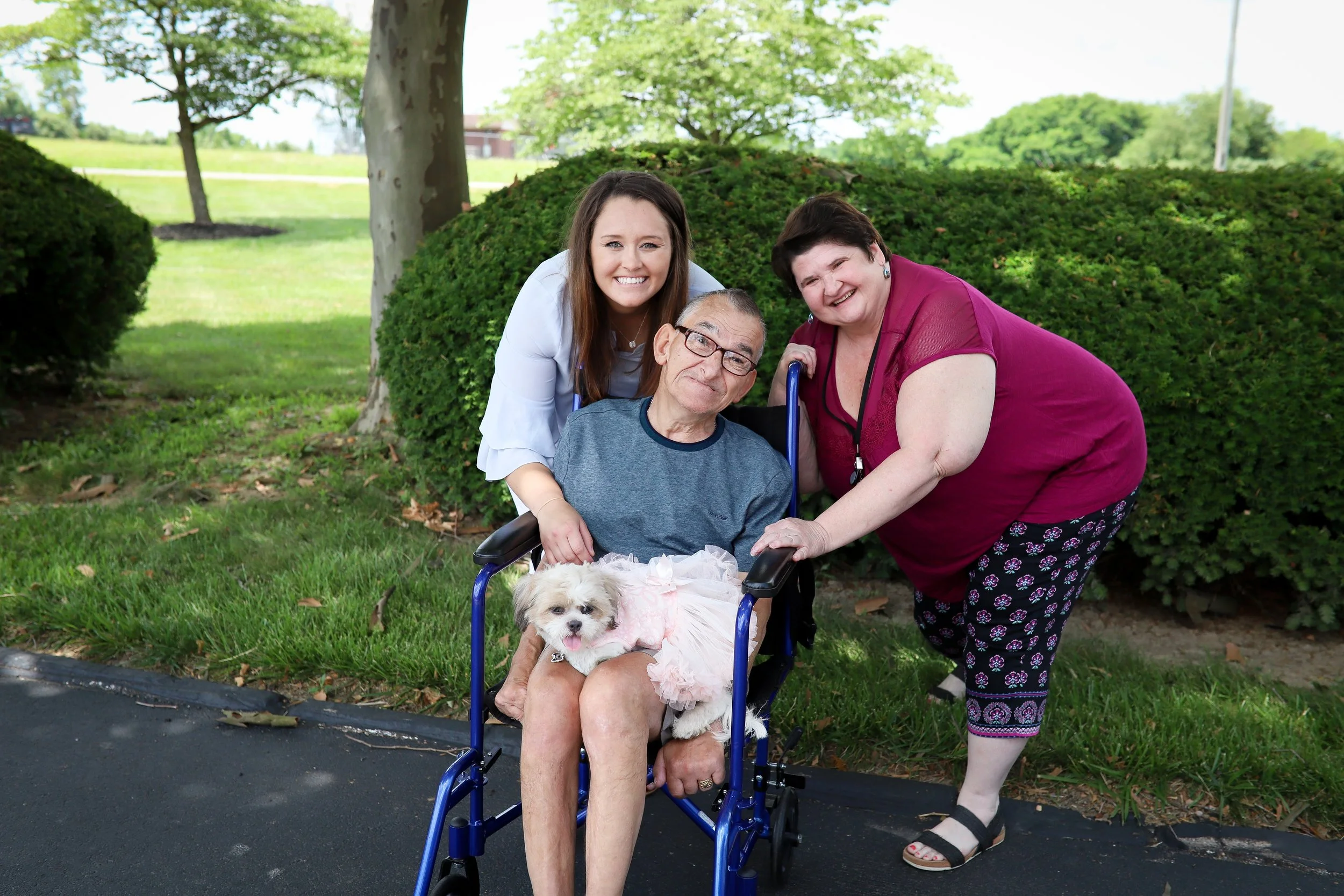 elderly man with disability sitting in wheel chair with two women standing on each side of him.  all three smiling at camera