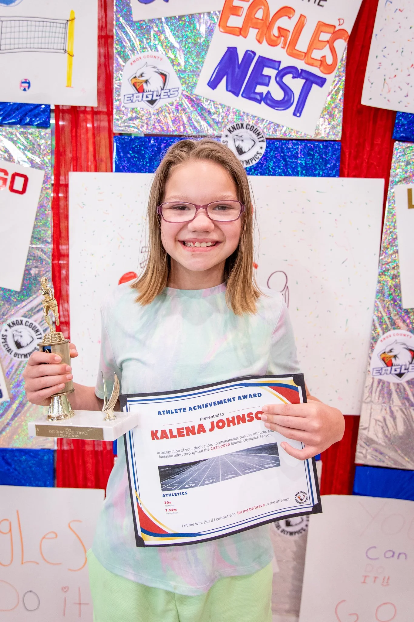 portrait of a person standing in front of a special olympics back drop, holding their trophy and certificate