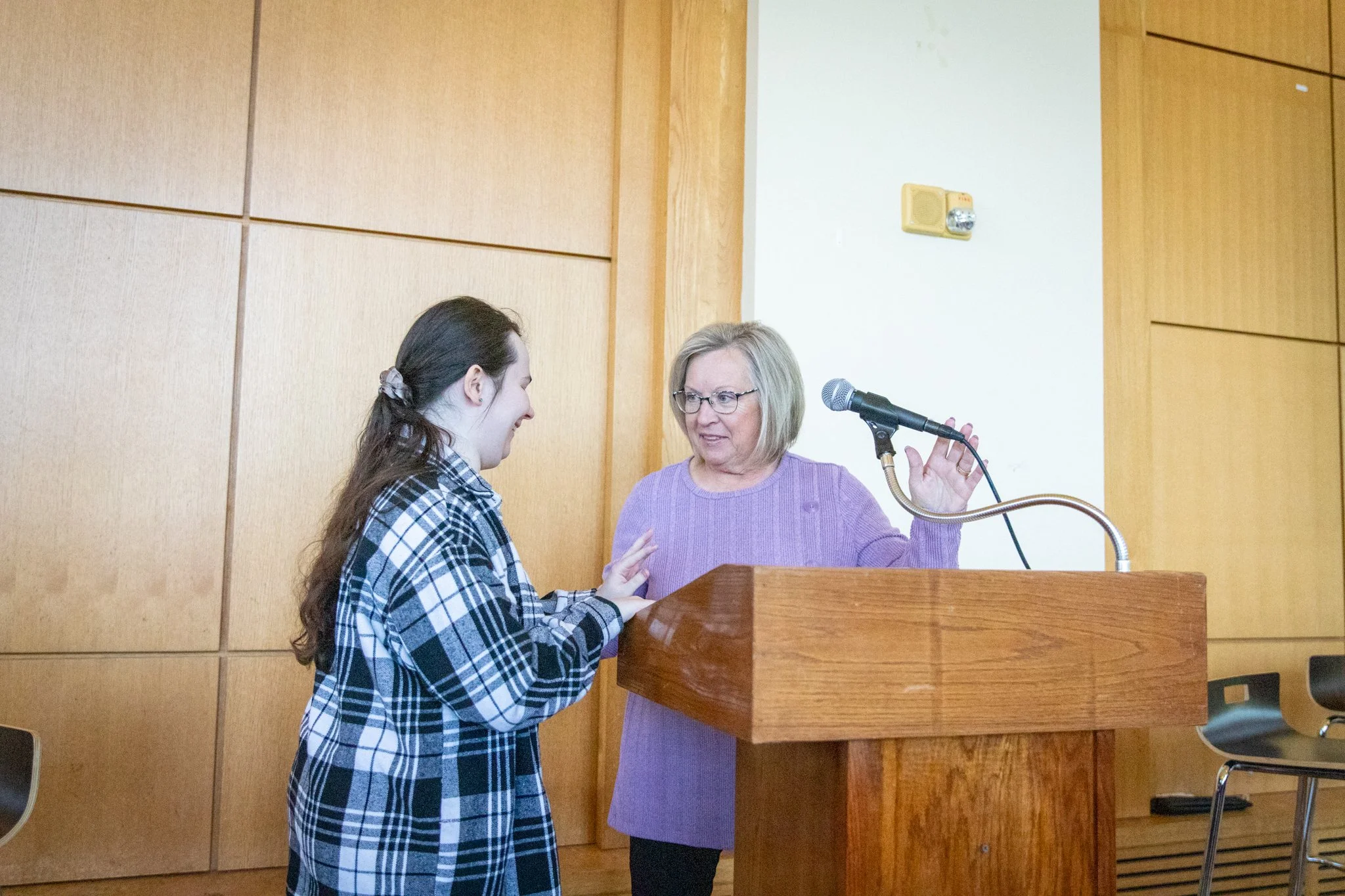 Person at a podium adjusts a microphone while speaking with another person standing beside them.