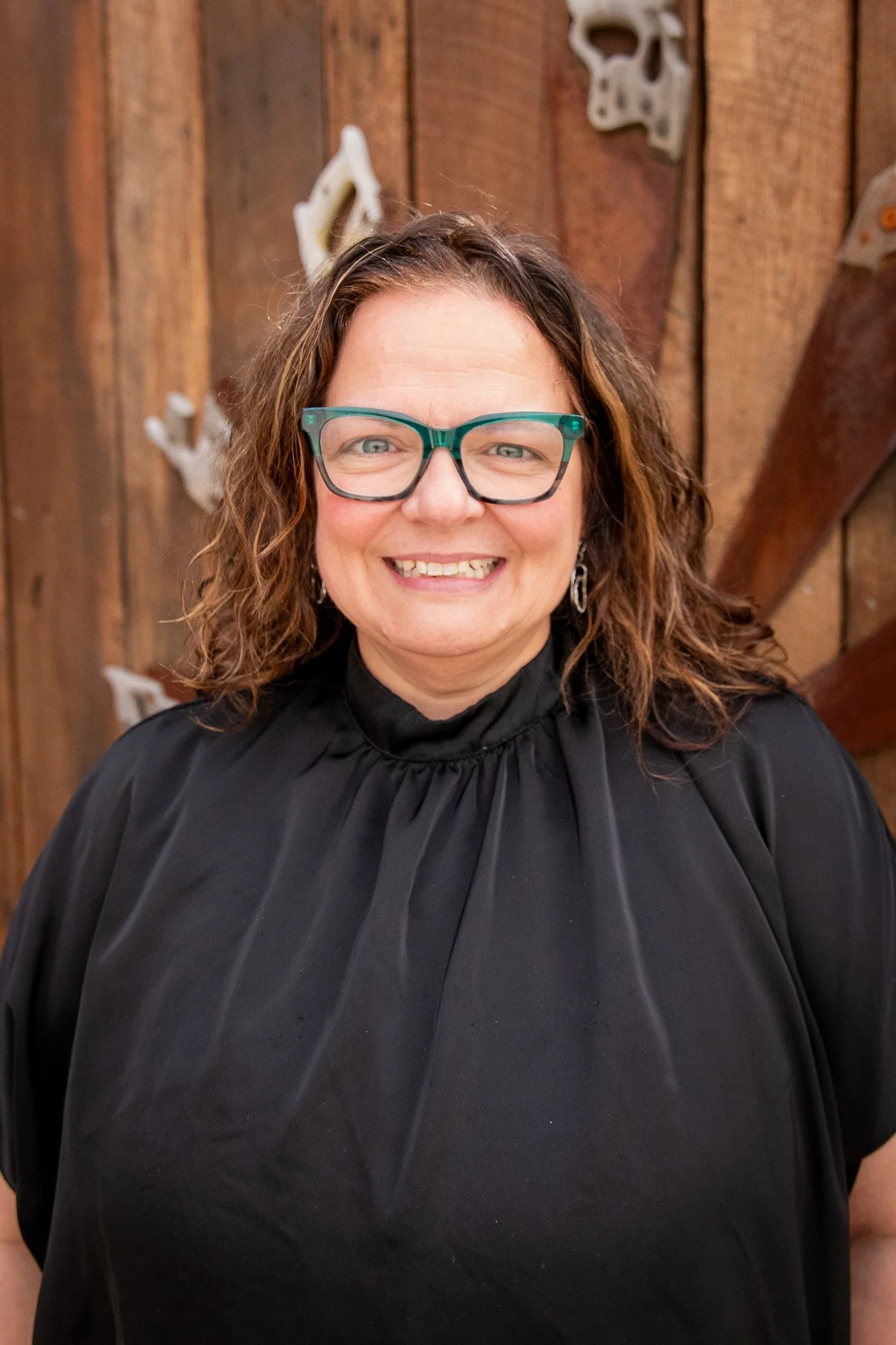 Close-up outdoor portrait of a person with shoulder-length wavy hair wearing green-framed glasses and a black high-neck top. The person is standing in front of a wooden fence with decorative metal accents, with a softly blurred background.
