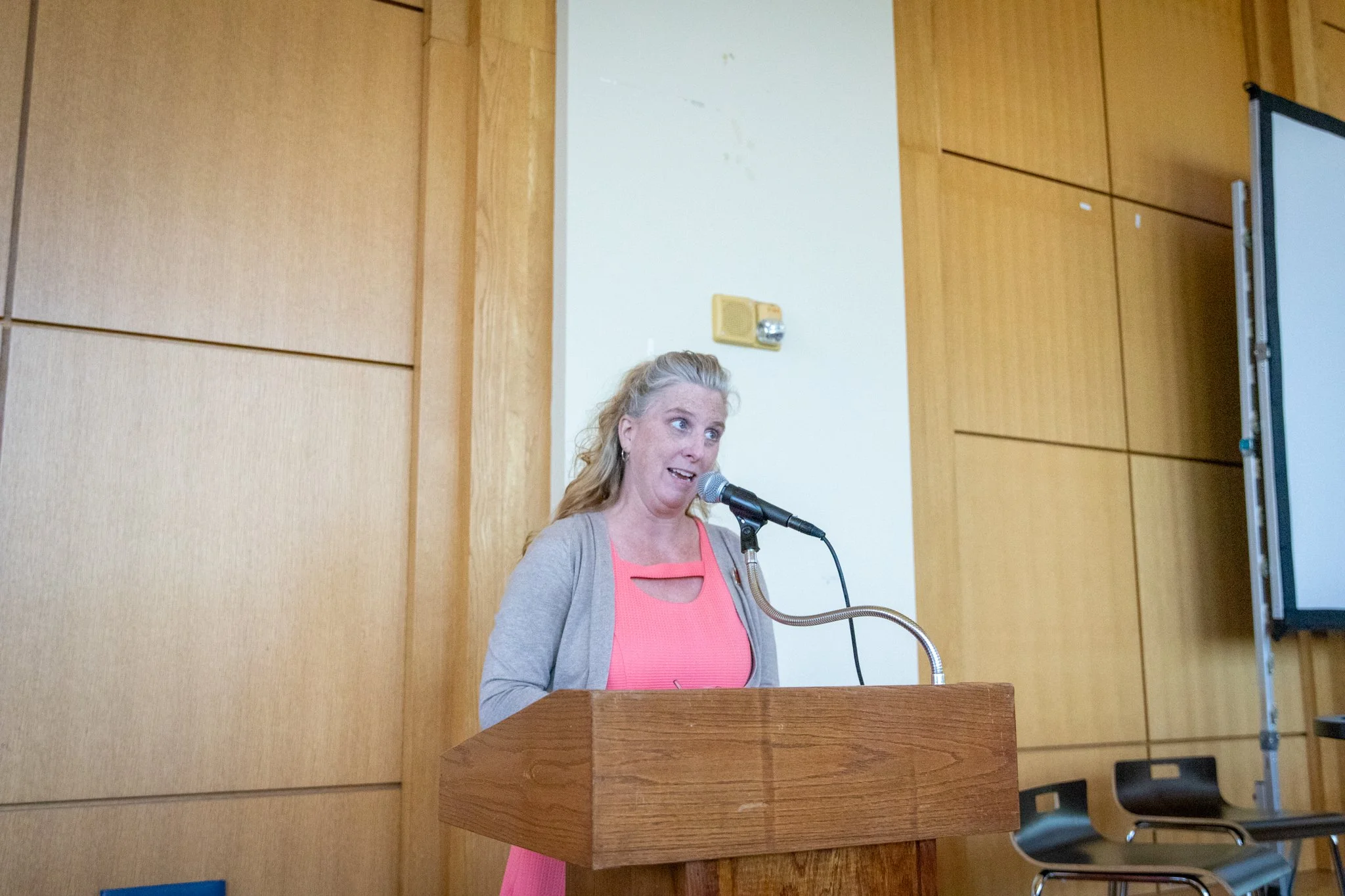 Person stands at a wooden podium, speaking into a microphone in a wood-paneled room.