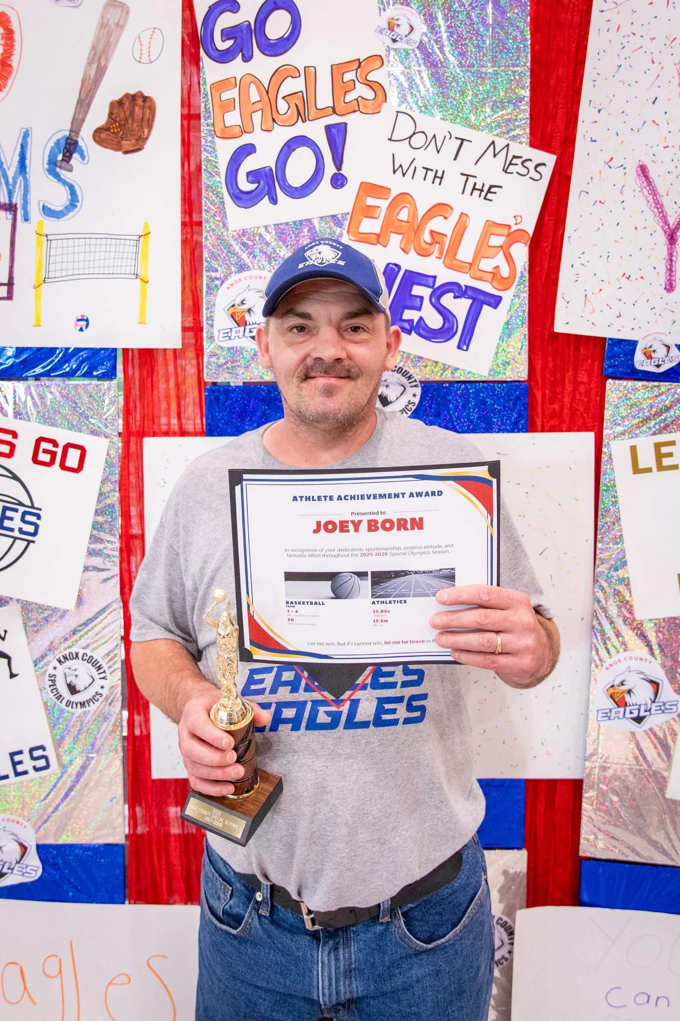 portrait of a person standing in front of a special olympics back drop, holding their trophy and certificate