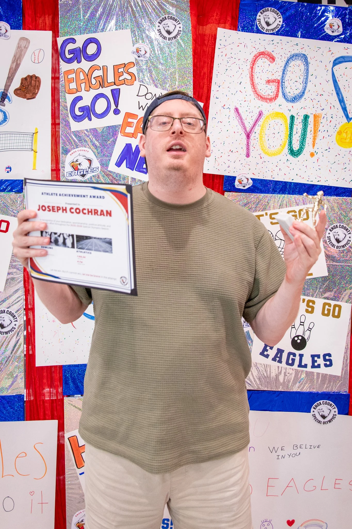 portrait of a person standing in front of a special olympics back drop, holding their trophy and certificate
