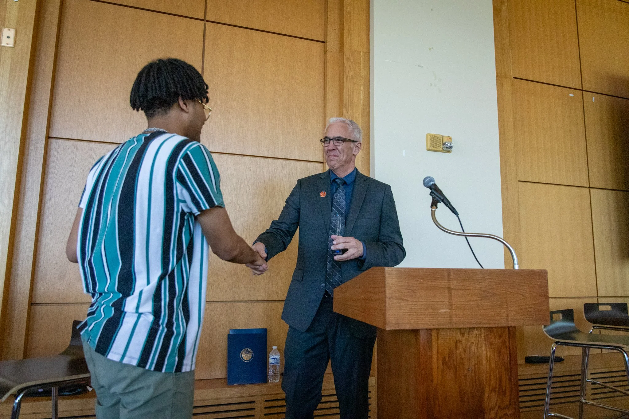 person in a suit shakes hands with a young man in a striped shirt at a podium