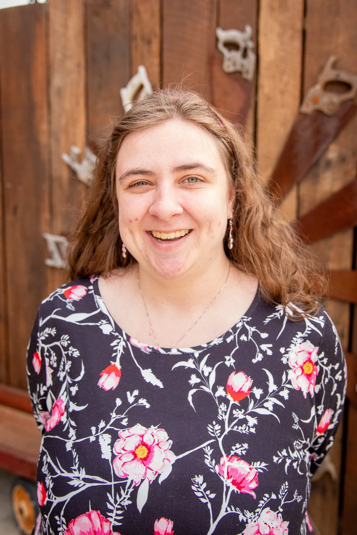 Close-up outdoor portrait of a person with long hair wearing a black top with a pink and white floral pattern. The person is standing in front of a wooden fence decorated with metal tools, with a softly blurred background.