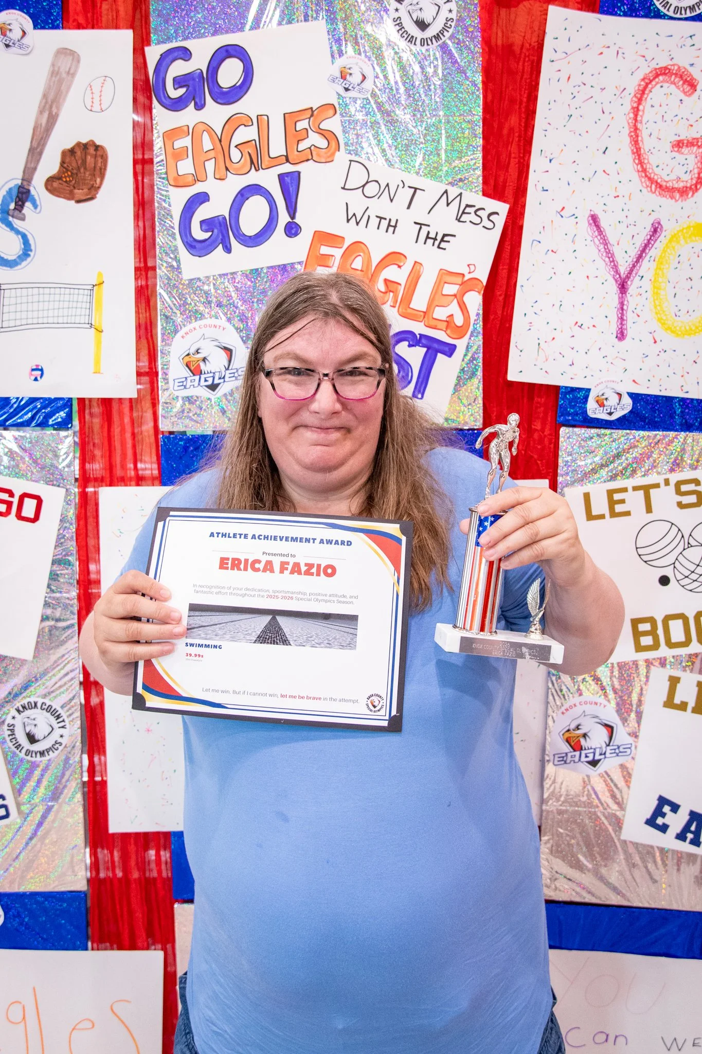 portrait of a person standing in front of a special olympics back drop, holding their trophy and certificate
