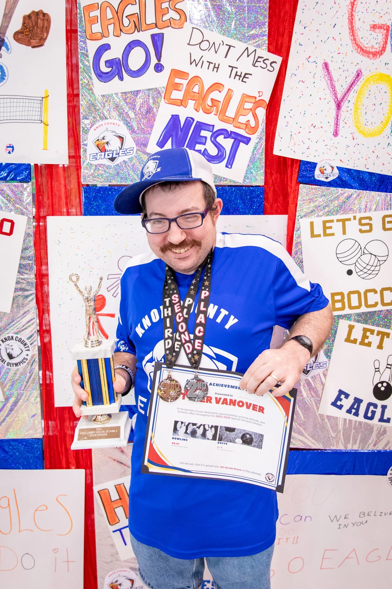 portrait of a person standing in front of a special olympics back drop, holding their trophy and certificate