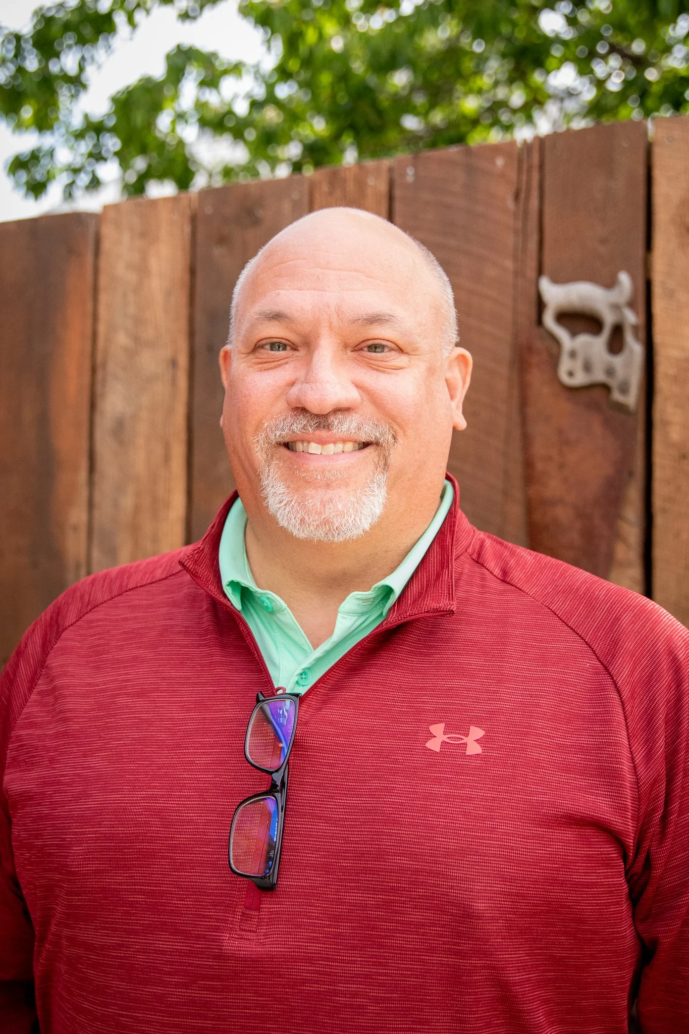 Vertical, full color image. portrait of a white man who is looking directly at the camera and smiling.  He has short grey hair.  He is wearing a mint green polo and a maroon red pullover on top.  there is a rustic wood fence in the background.