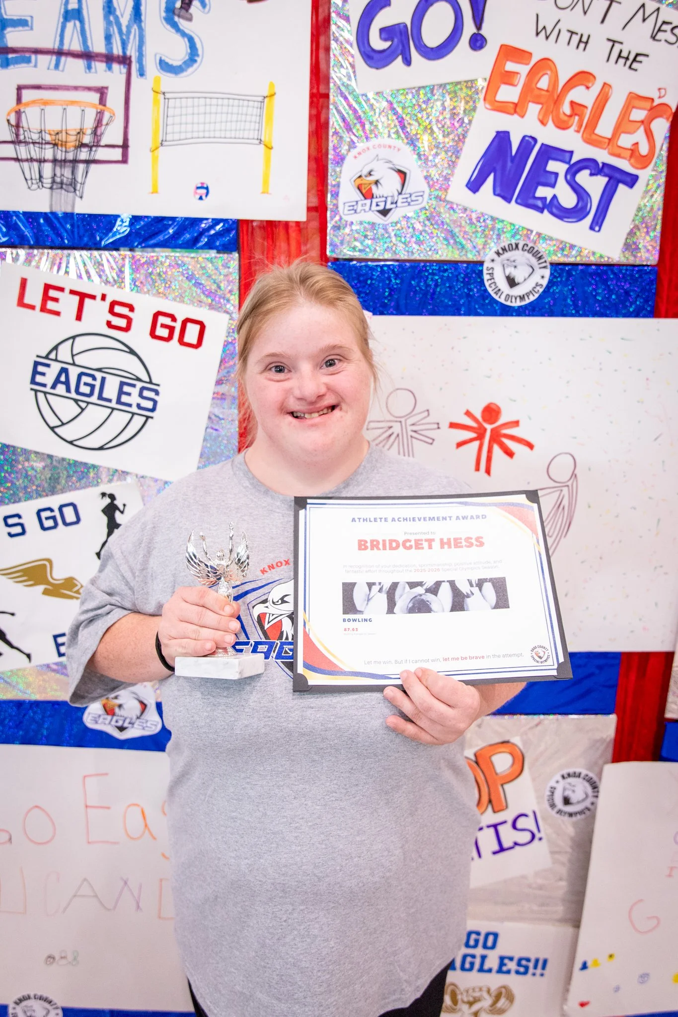 portrait of a person standing in front of a special olympics back drop, holding their trophy and certificate