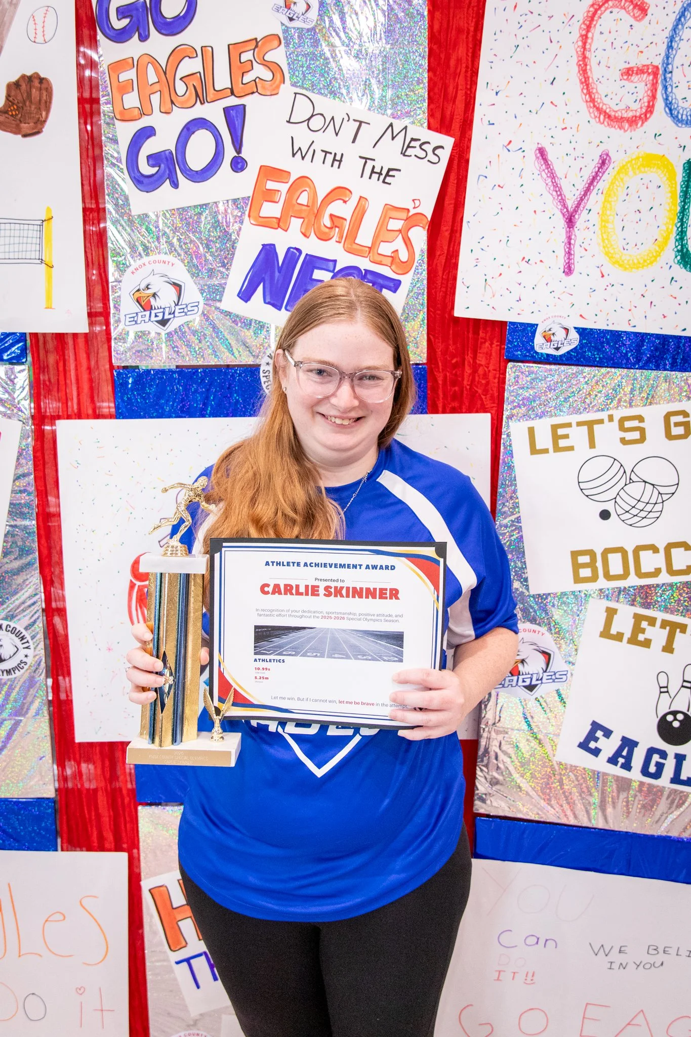 portrait of a person standing in front of a special olympics back drop, holding their trophy and certificate