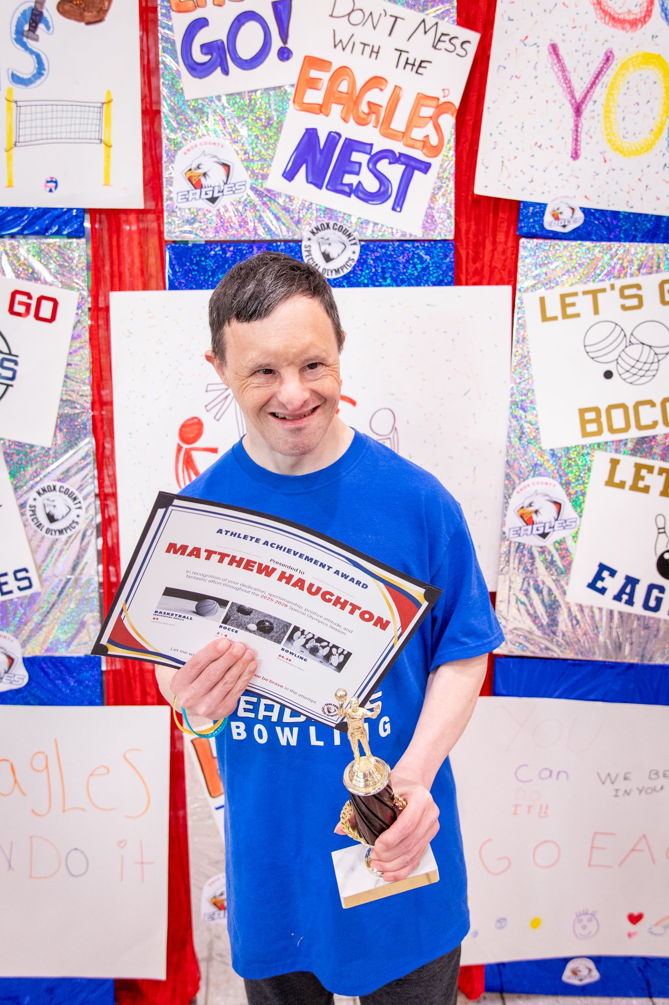 portrait of a person standing in front of a special olympics back drop, holding their trophy and certificate