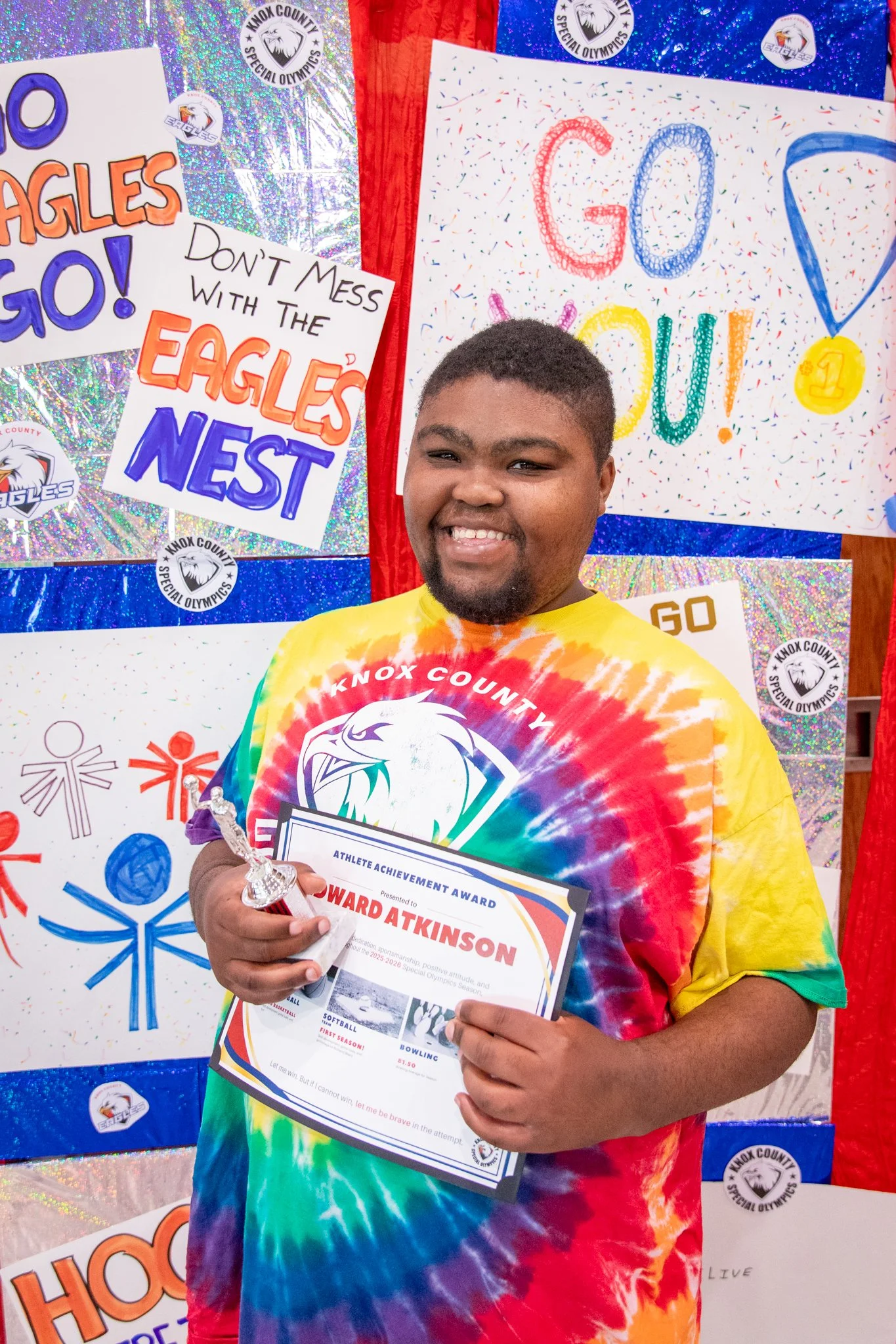 portrait of a person standing in front of a special olympics back drop, holding their trophy and certificate