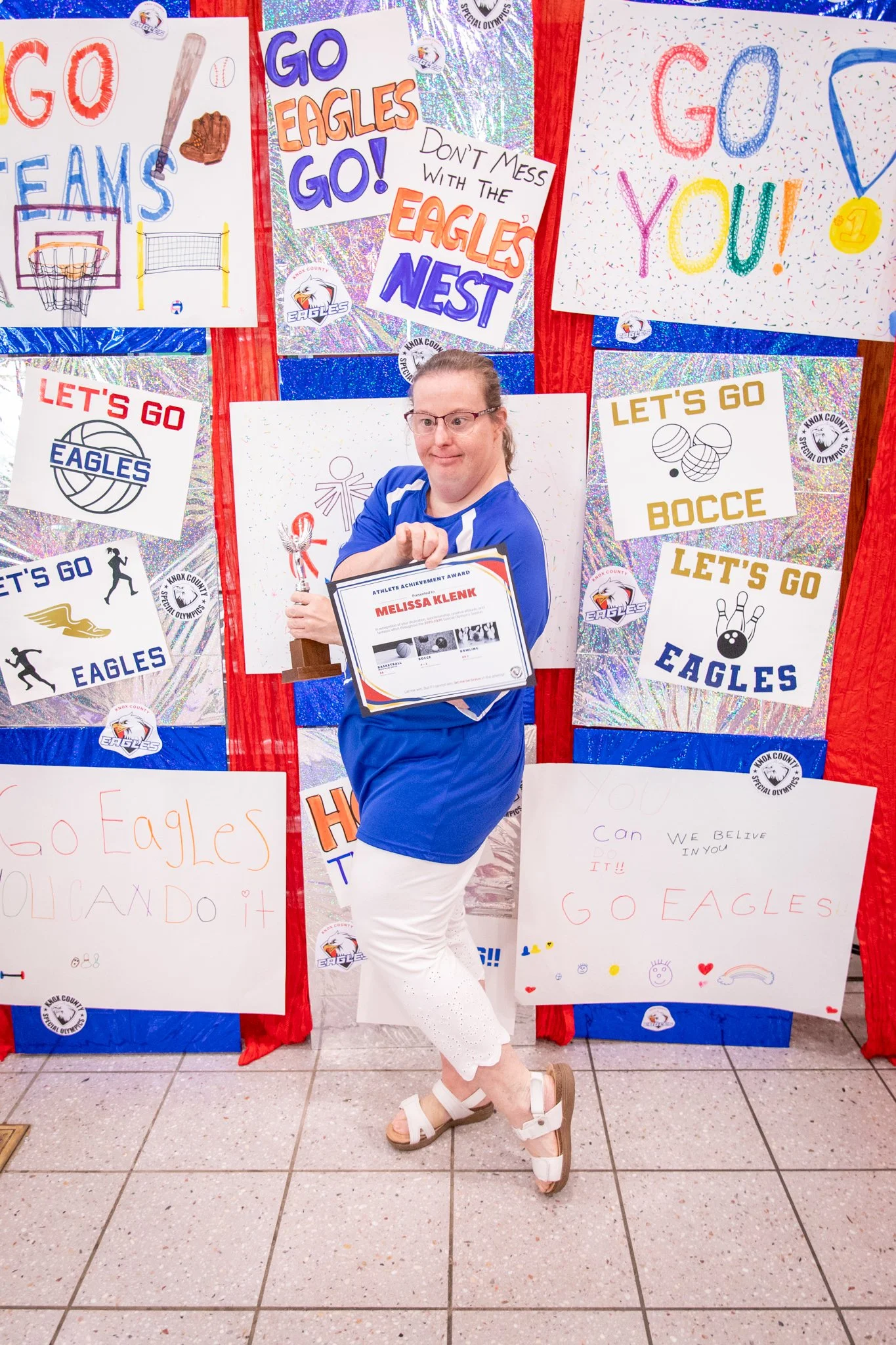 portrait of a person standing in front of a special olympics back drop, holding their trophy and certificate