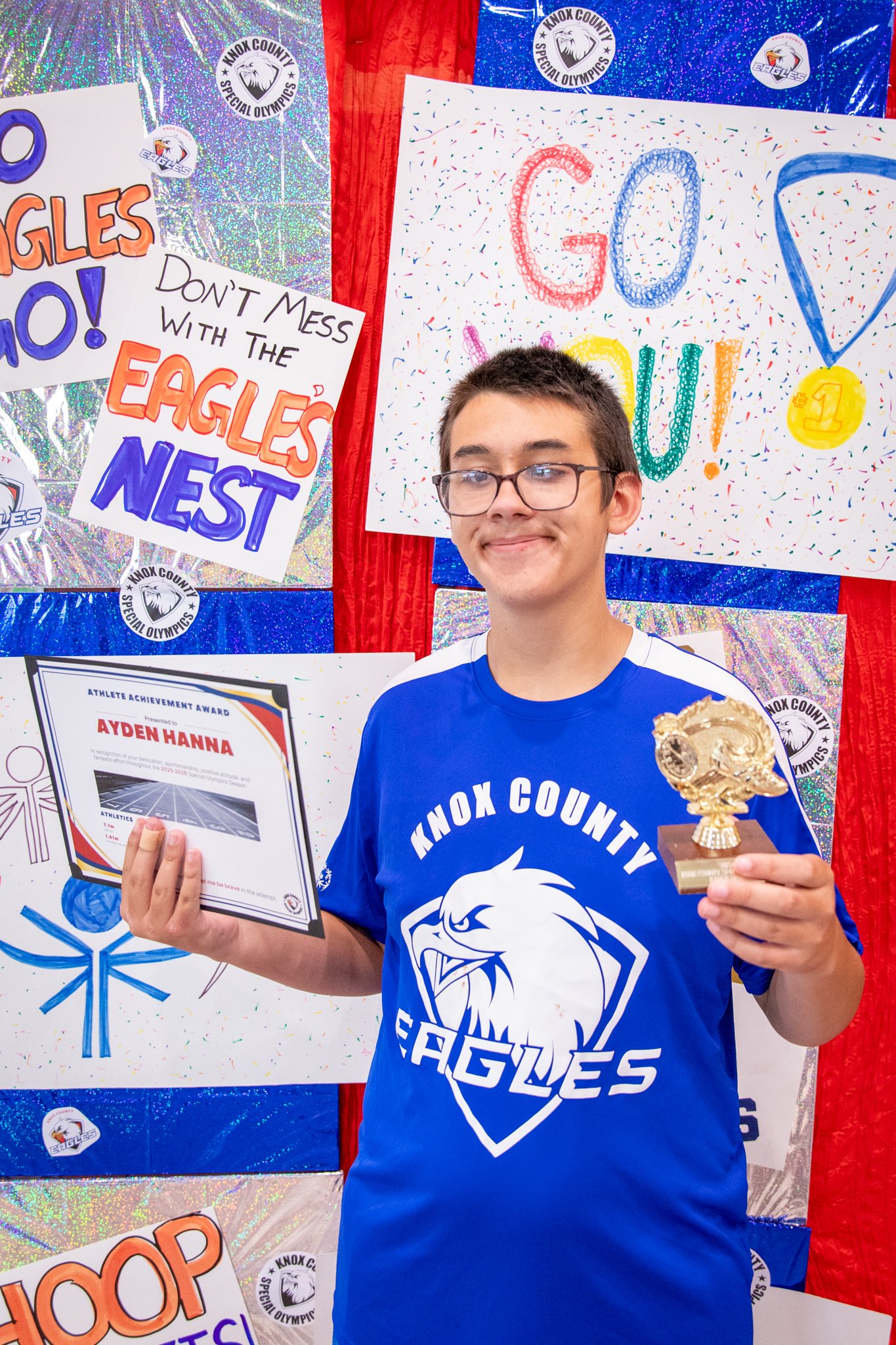 portrait of a person standing in front of a special olympics back drop, holding their trophy and certificate