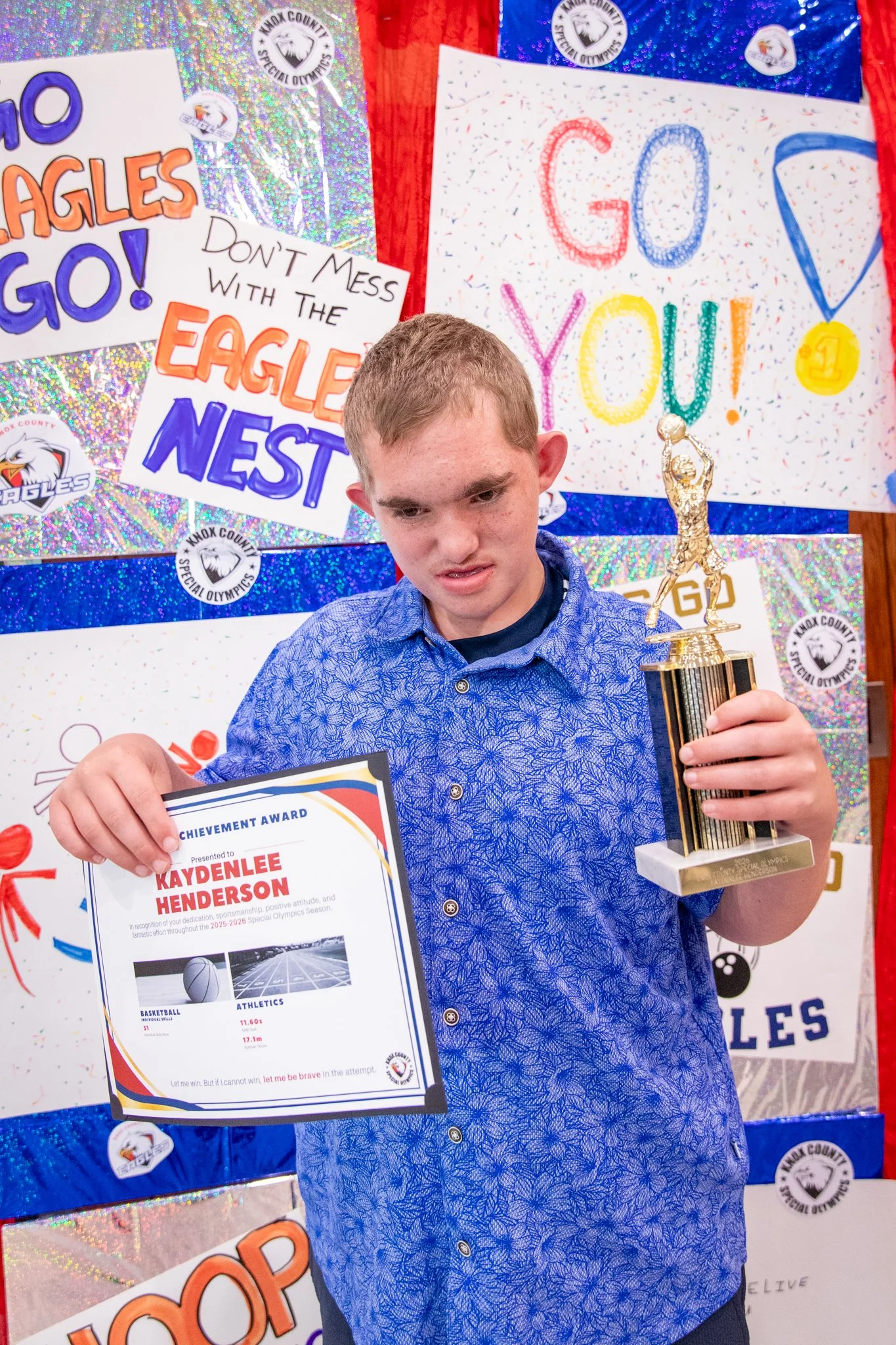 portrait of a person standing in front of a special olympics back drop, holding their trophy and certificate