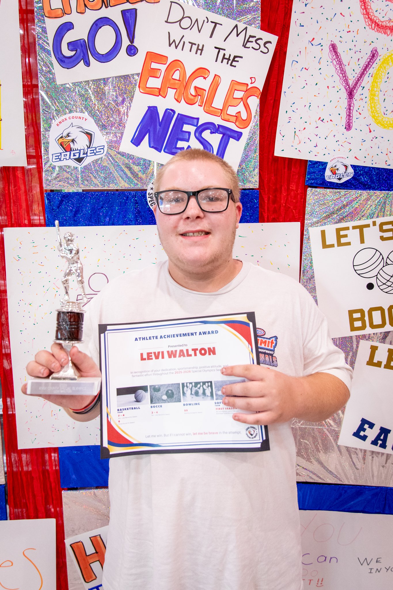 portrait of a person standing in front of a special olympics back drop, holding their trophy and certificate