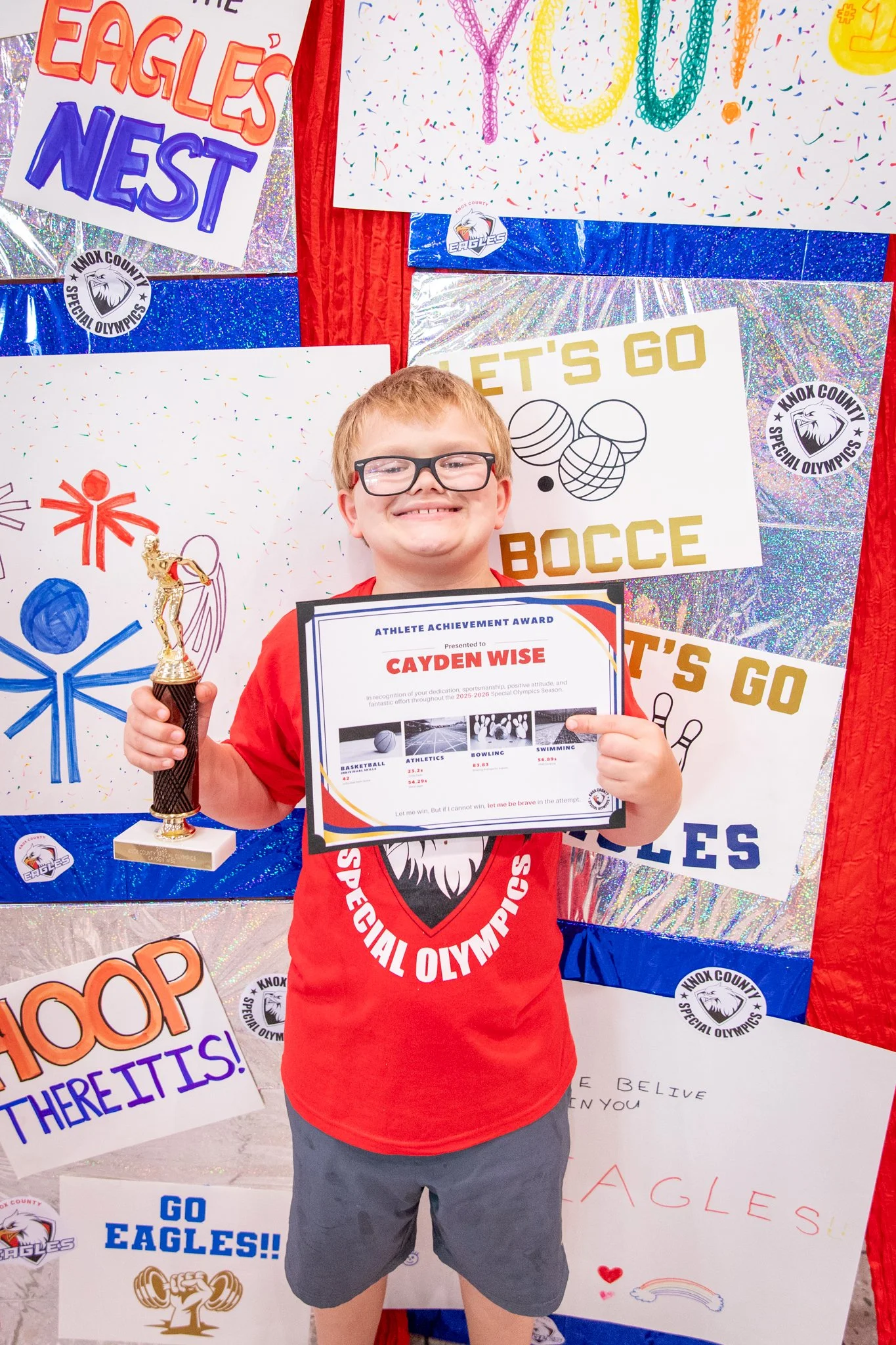 portrait of a person standing in front of a special olympics back drop, holding their trophy and certificate