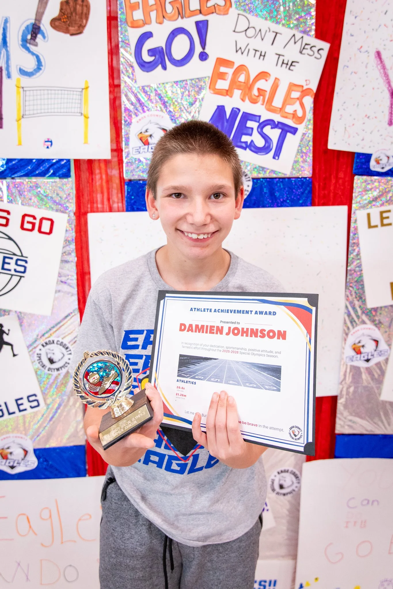 portrait of a person standing in front of a special olympics back drop, holding their trophy and certificate