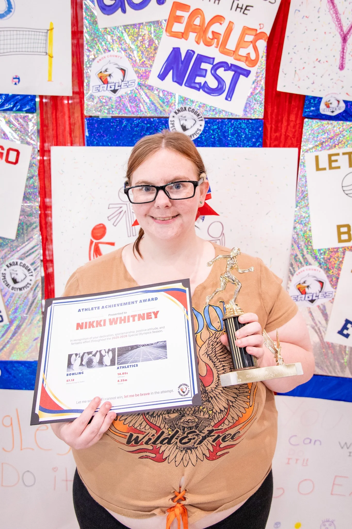 portrait of a person standing in front of a special olympics back drop, holding their trophy and certificate
