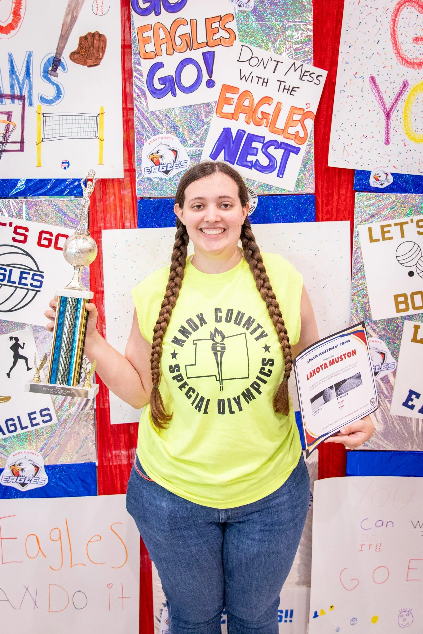 portrait of a person standing in front of a special olympics back drop, holding their trophy and certificate