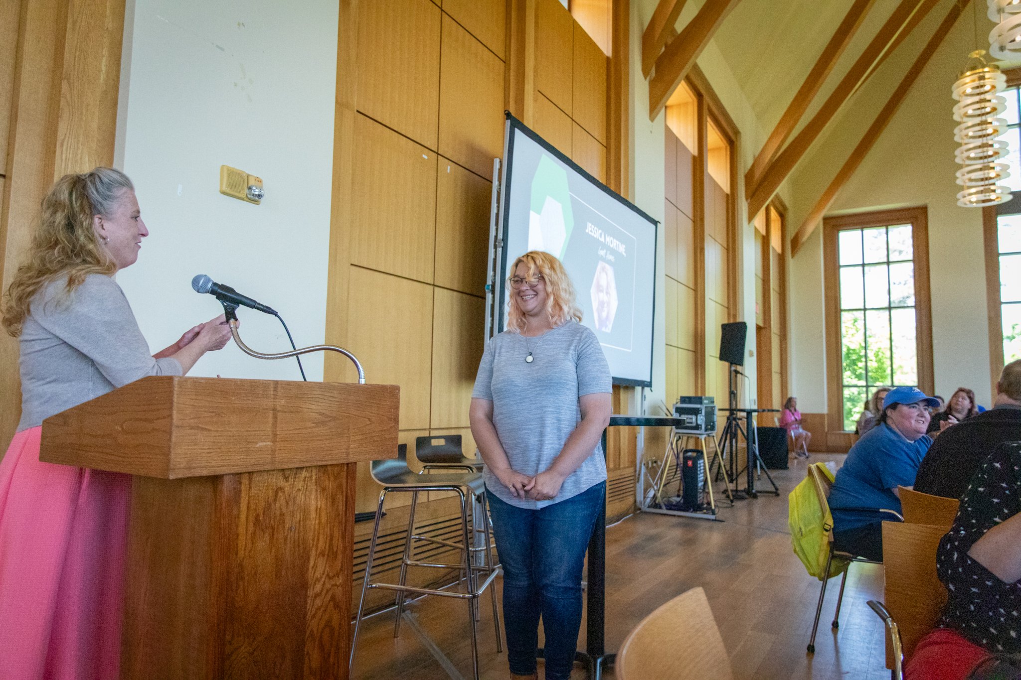 Person at a podium gestures toward another person standing nearby during a presentation in a bright event space.