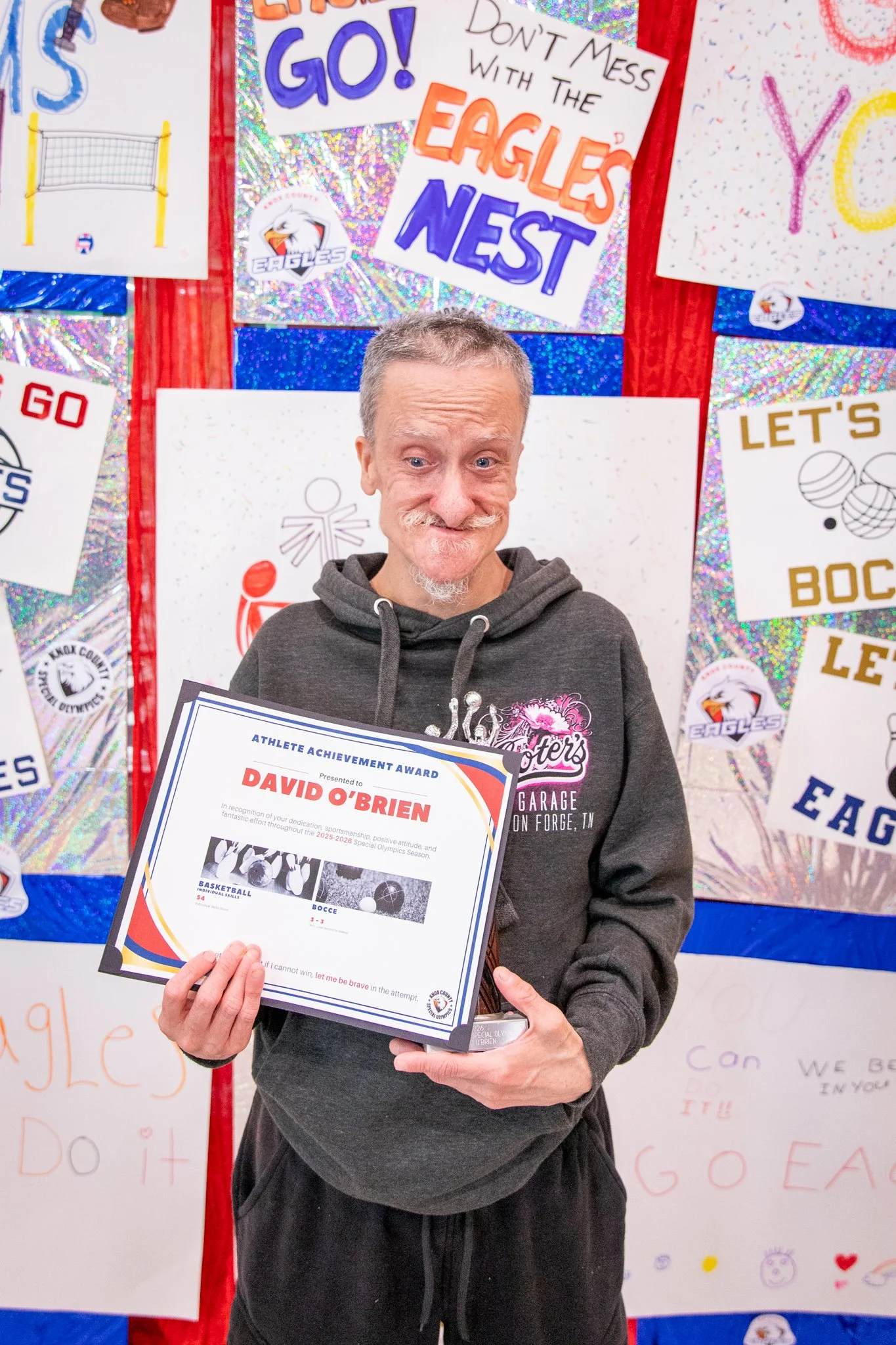 portrait of a person standing in front of a special olympics back drop, holding their trophy and certificate