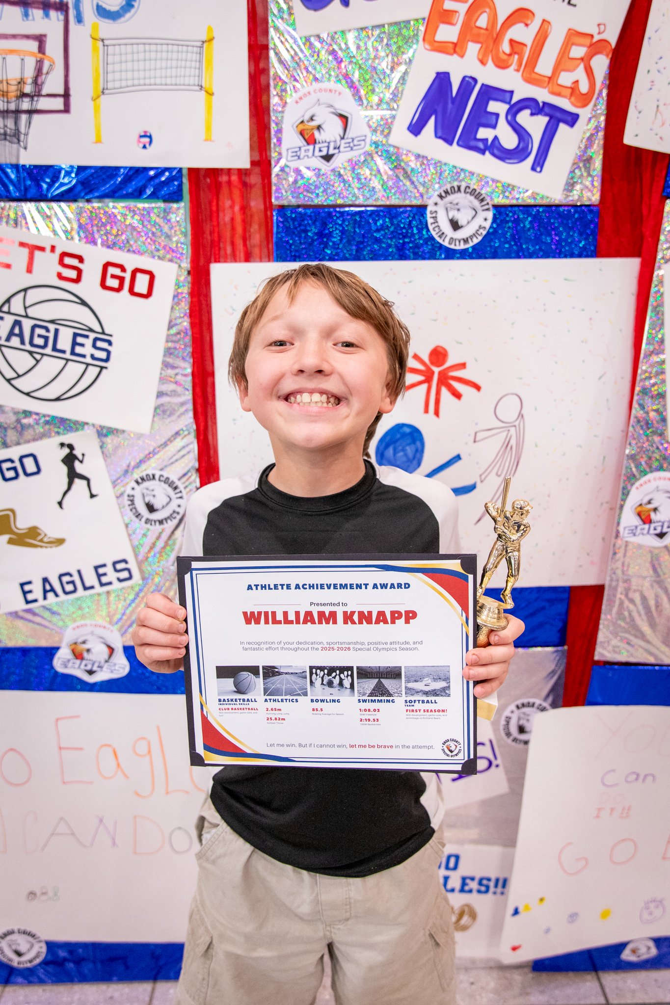 portrait of a person standing in front of a special olympics back drop, holding their trophy and certificate