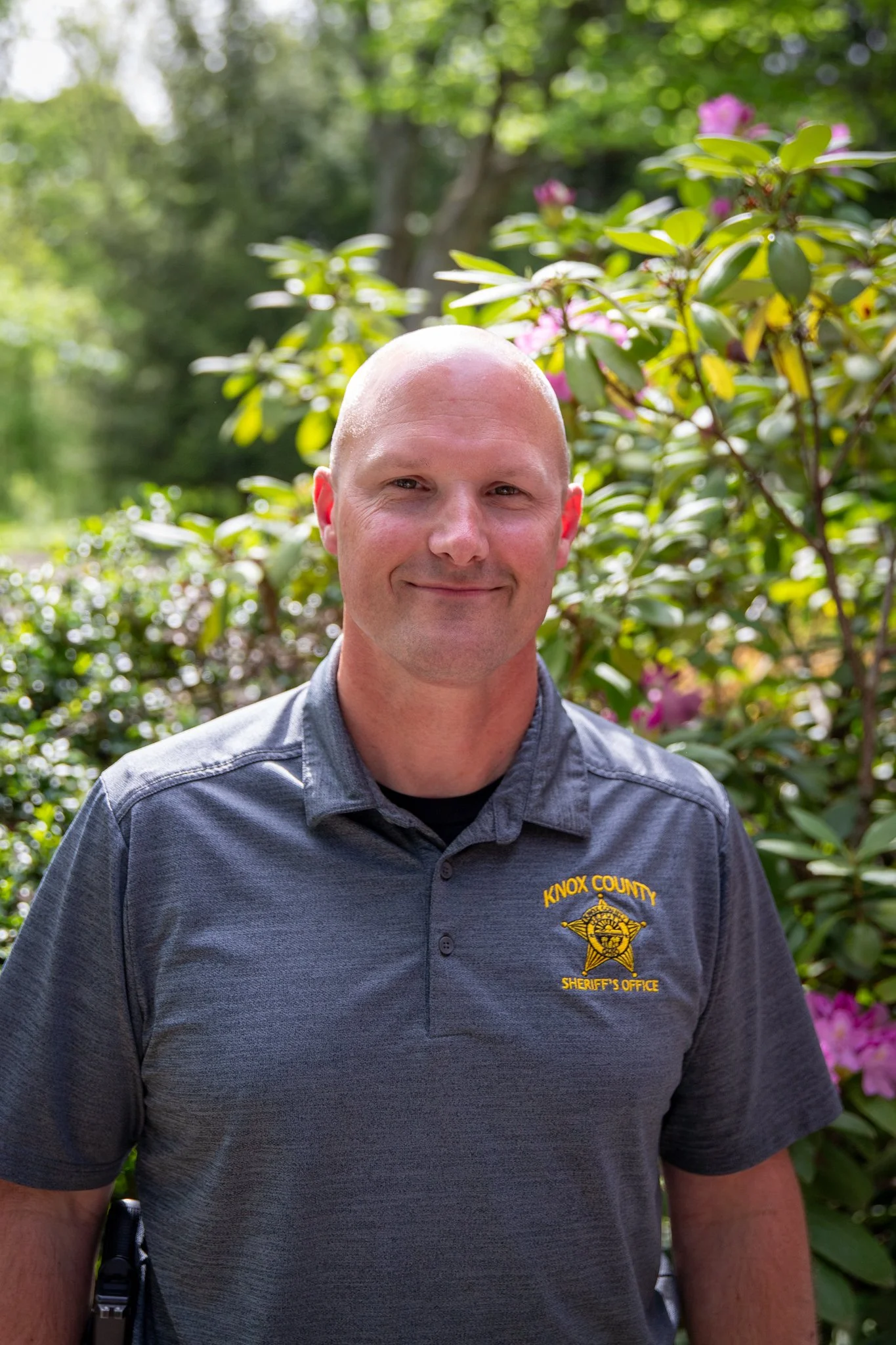 close up outdoor portrait of a person with a shaved head wearing a grey polo shirt with a yellow "Knox County Sheriff's Office" emblem on the chest.  The person is standing in front of green shrubs and flowering plants, with blurred background