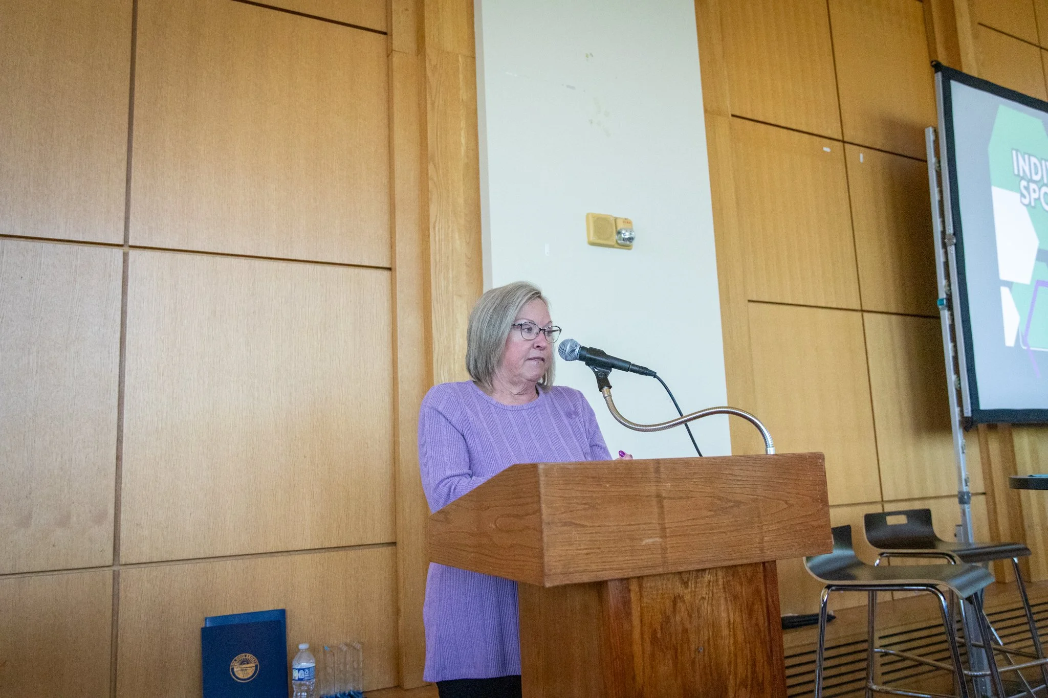 Person stands at a wooden podium speaking into a microphone in a wood-paneled event space.