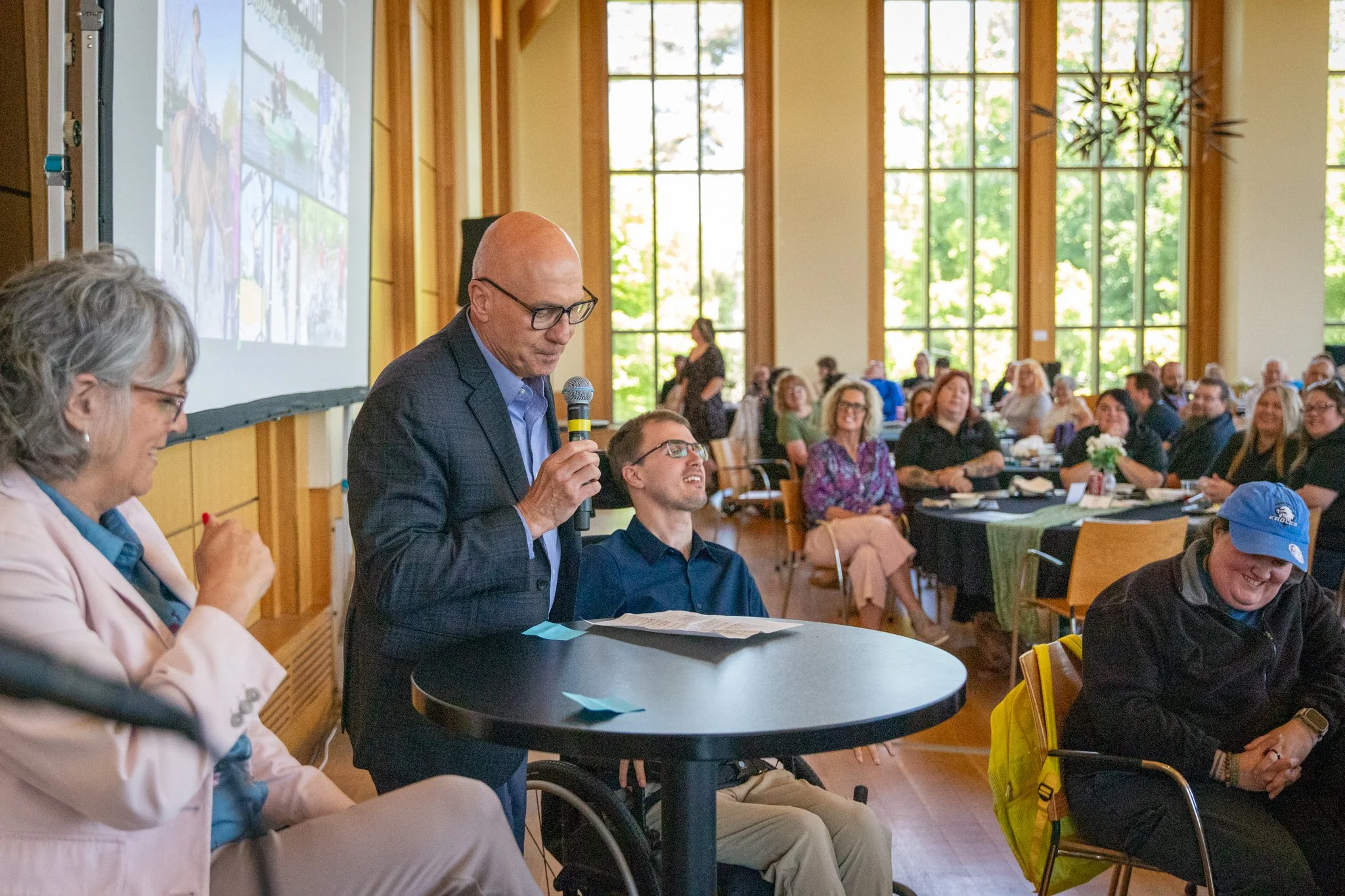 Person in a suit speaks into a microphone at a round table while audience members look on in a bright event space.