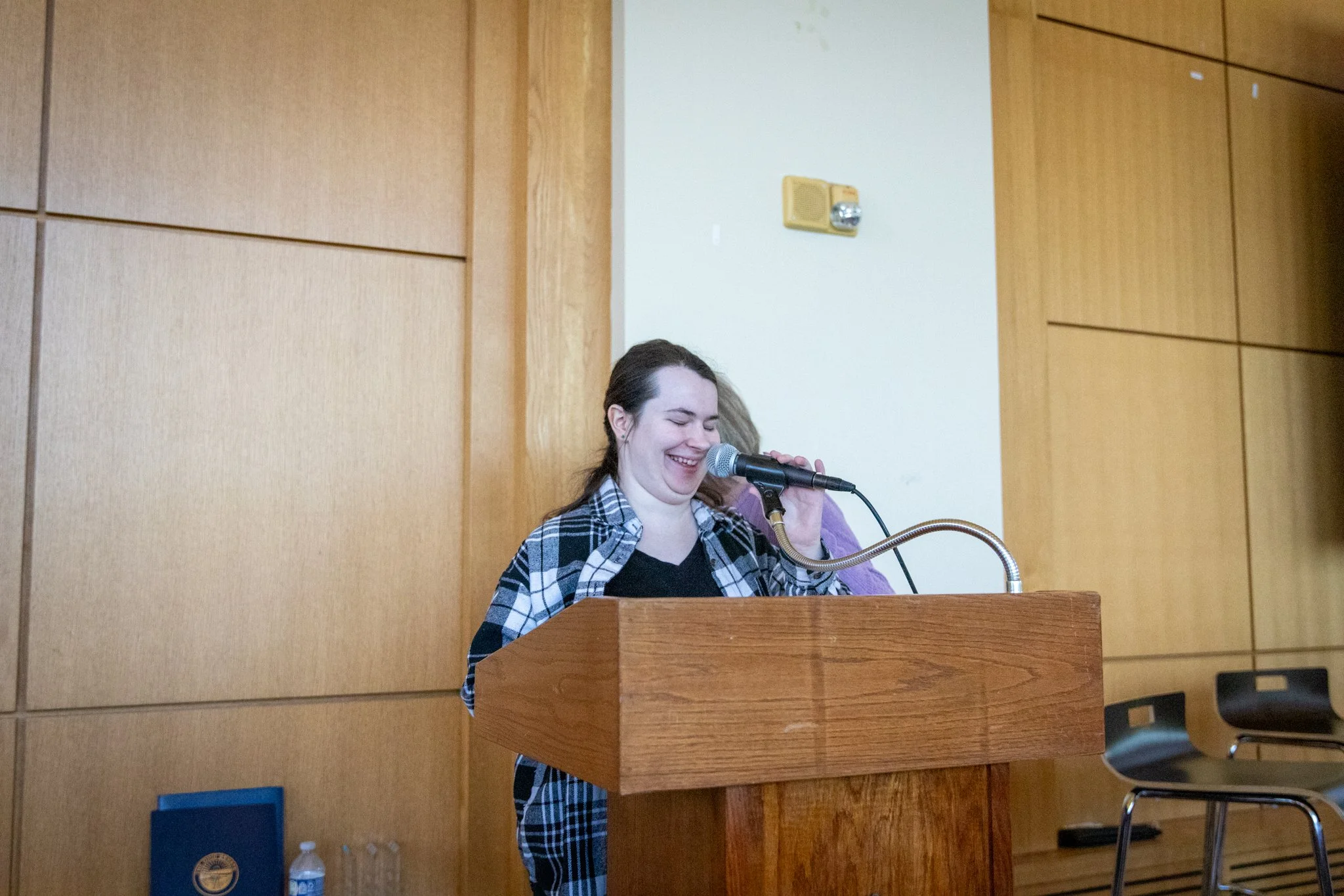 Person stands at a wooden podium, smiling while speaking into a microphone in a wood-paneled room.