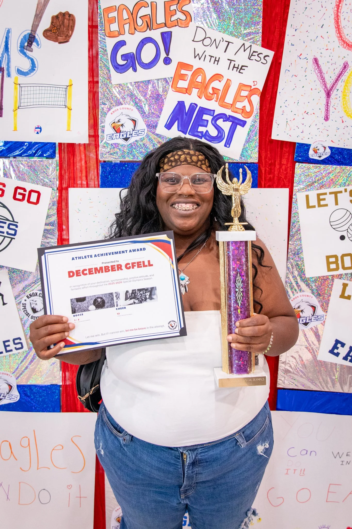 portrait of a person standing in front of a special olympics back drop, holding their trophy and certificate
