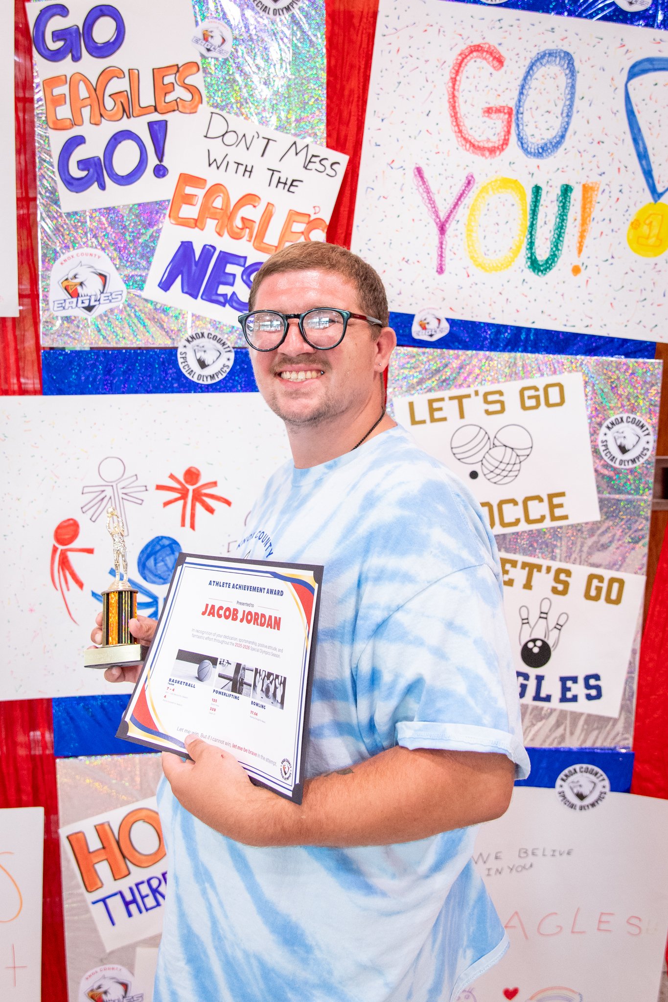 portrait of a person standing in front of a special olympics back drop, holding their trophy and certificate