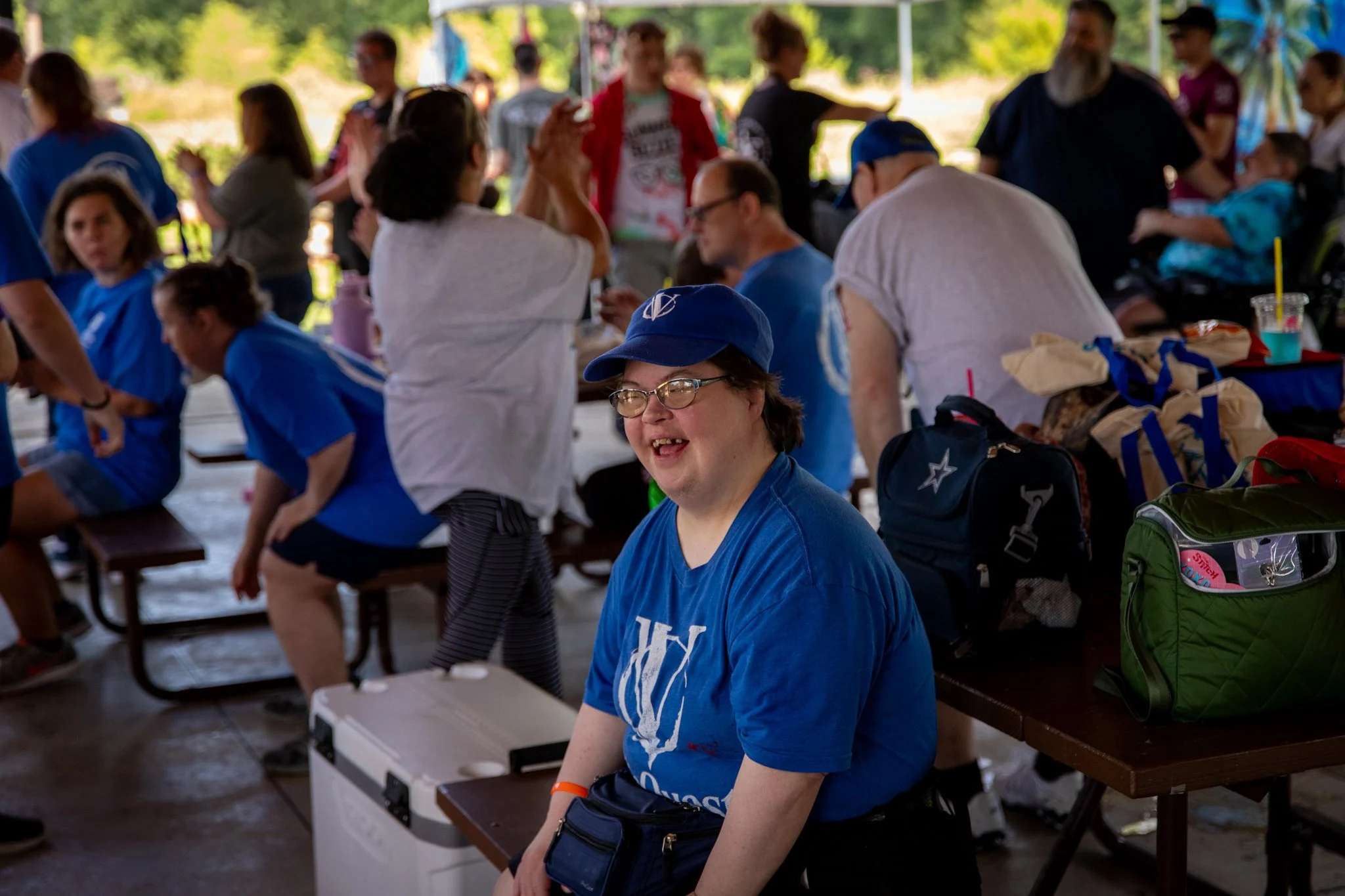 portrait of young woman with down syndrome wearing blue shirt and hat, smiling.  a large group of people are in the background