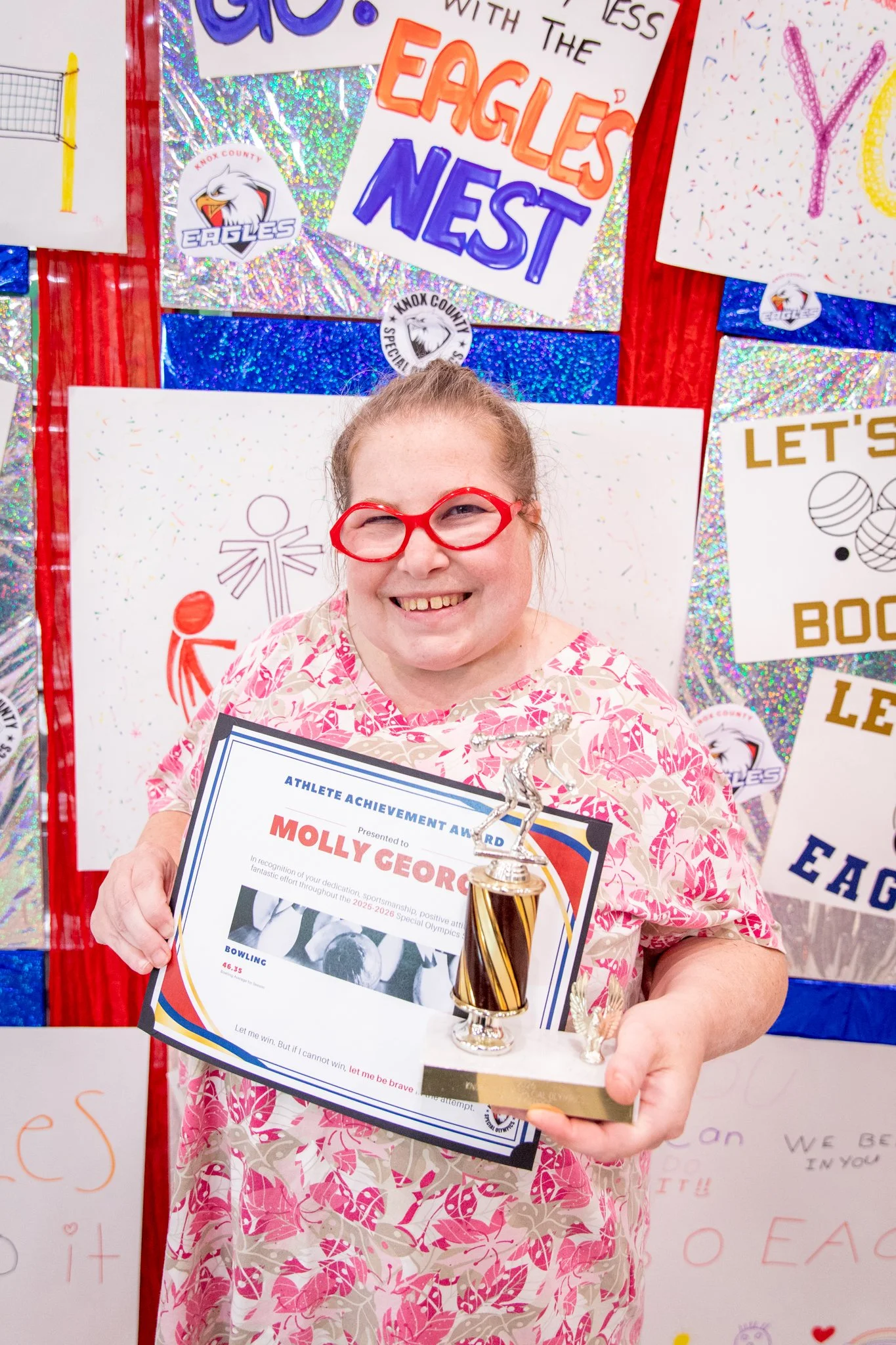 portrait of a person standing in front of a special olympics back drop, holding their trophy and certificate