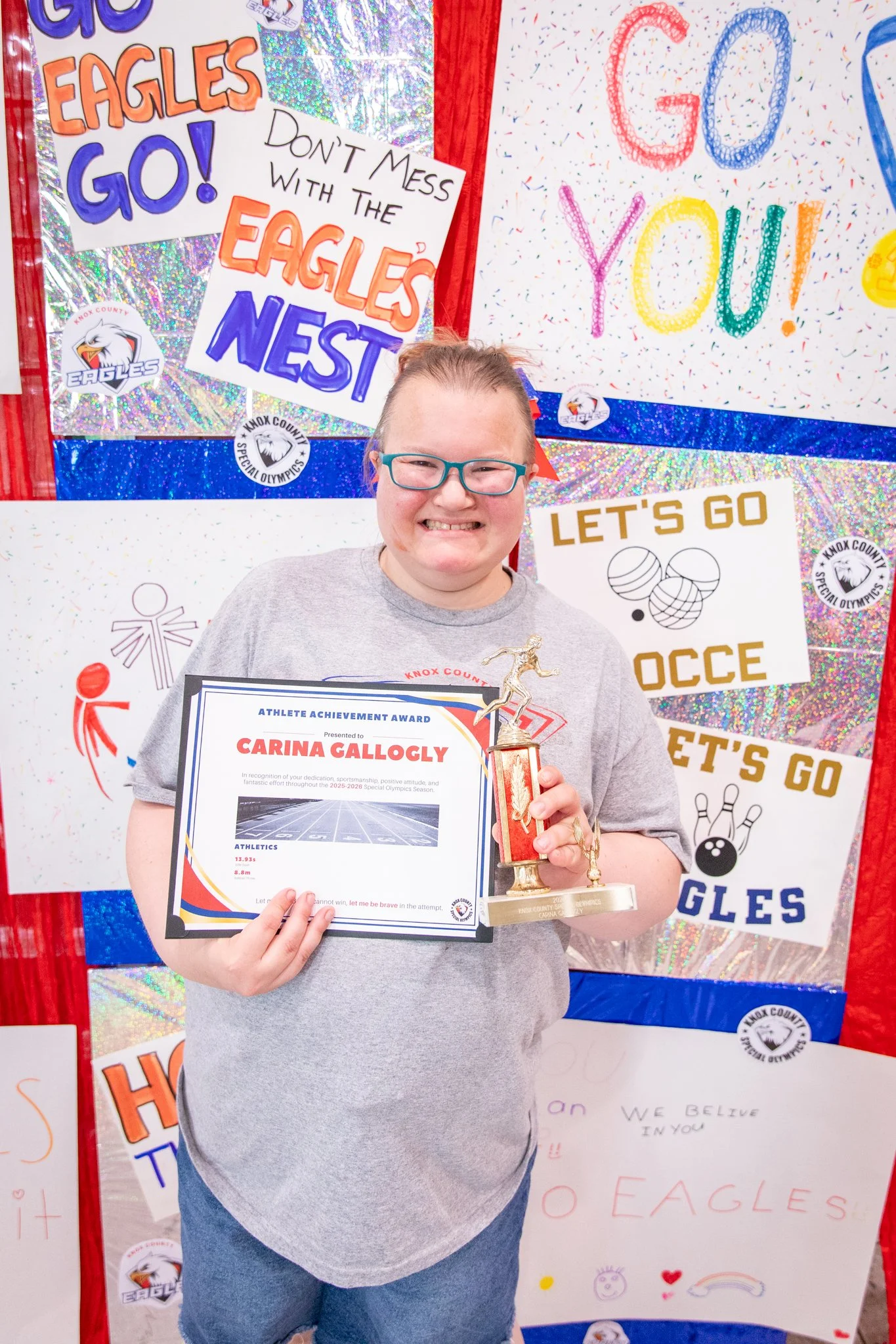 portrait of a person standing in front of a special olympics back drop, holding their trophy and certificate