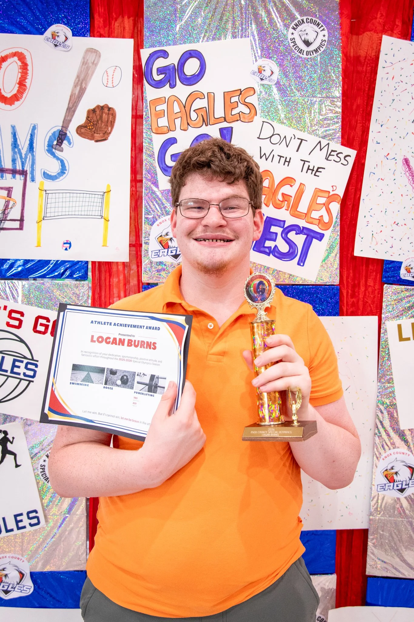 portrait of a person standing in front of a special olympics back drop, holding their trophy and certificate